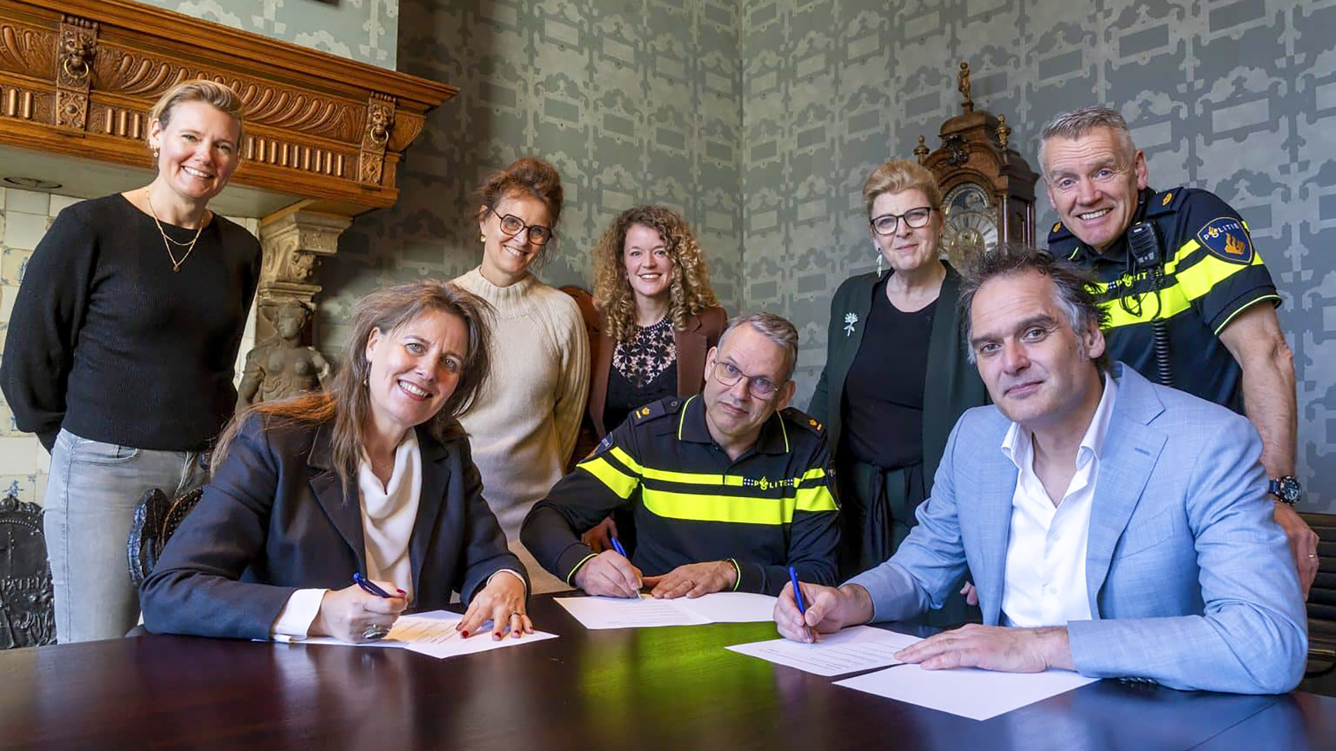 Groep mensen poseert in een kamer met een open haard; drie personen tekenen een document.