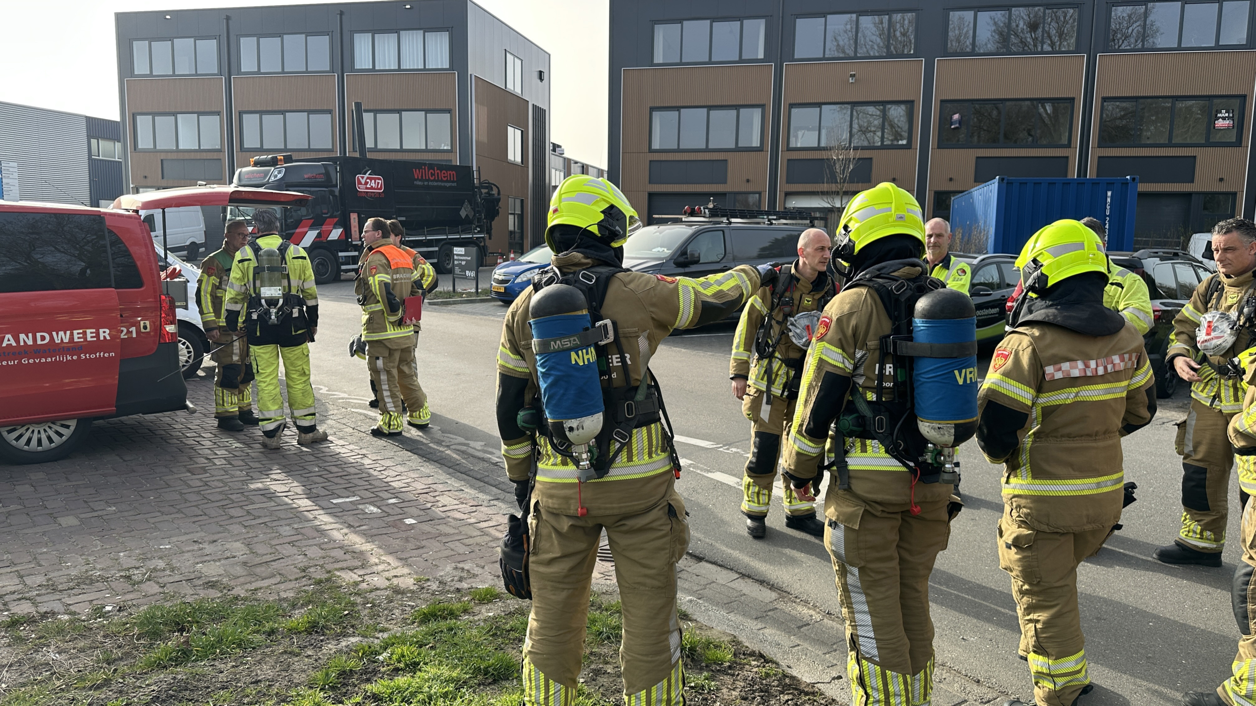Brandweerlieden in uniform overleggen bij een rode brandweerauto op straat, met panden op de achtergrond.