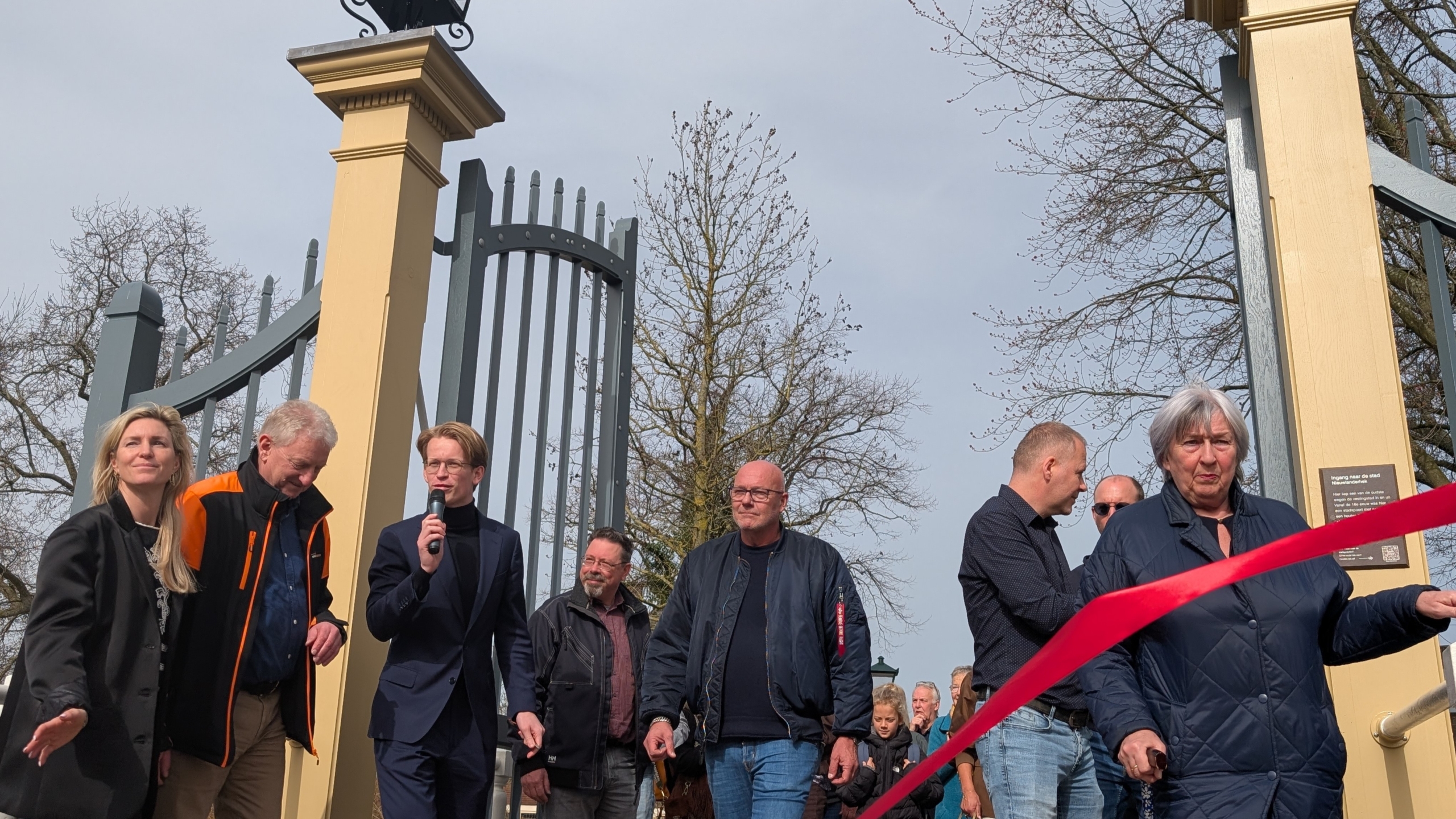 Mensen verzamelen zich bij een poort voor een ceremonie met een rood lint, een man houdt een microfoon vast.