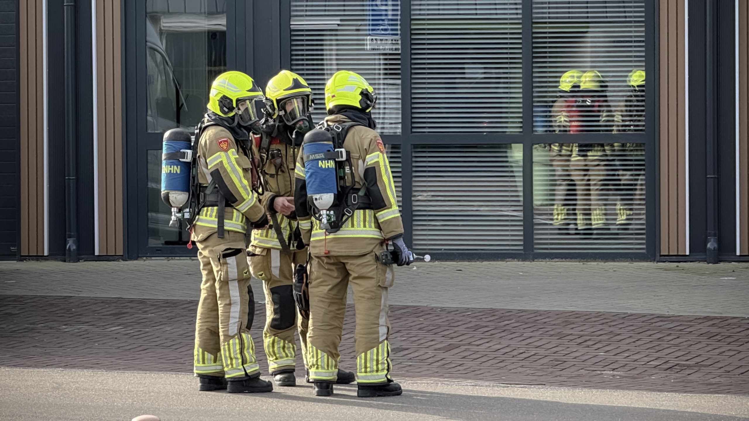 Brandweerlieden in beschermende kleding en helmen staan voor een gebouw met glazen gevel.