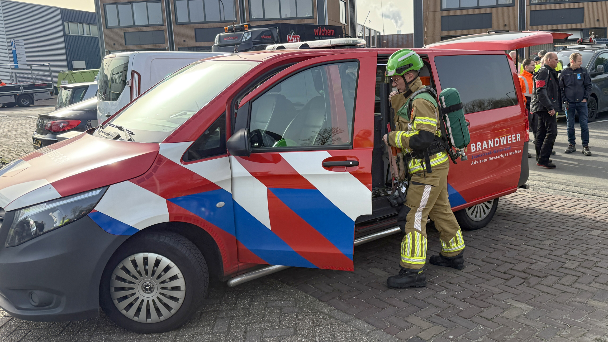 Brandweerman in uitrusting staat bij een brandweerwagen met geopende deur, omgeven door andere mensen en voertuigen.