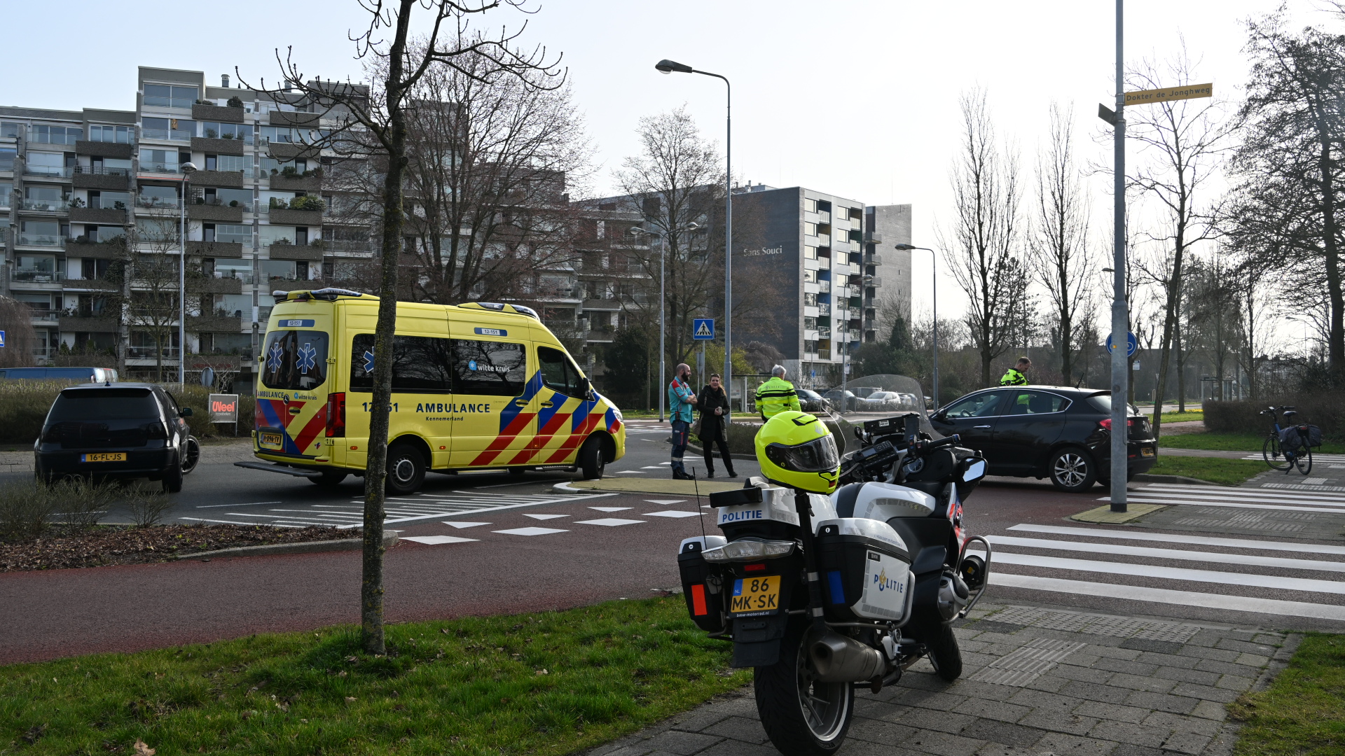 Gele ambulance en politie op motor bij een kruispunt naast flatgebouwen, met agenten en omstanders in gesprek.