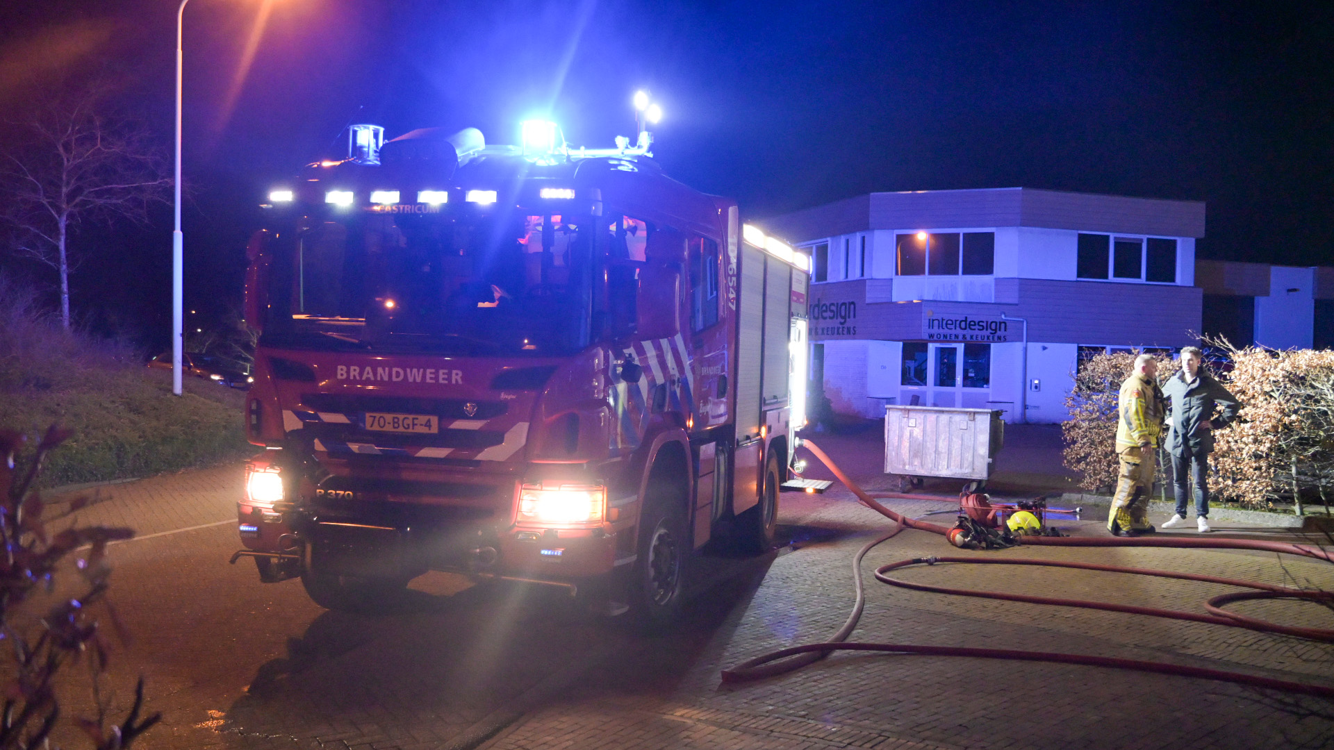 Brandweerwagen met felle lichten voor een gebouw op een verlichte straat; twee mannen staan naast uitgerolde brandslangen.