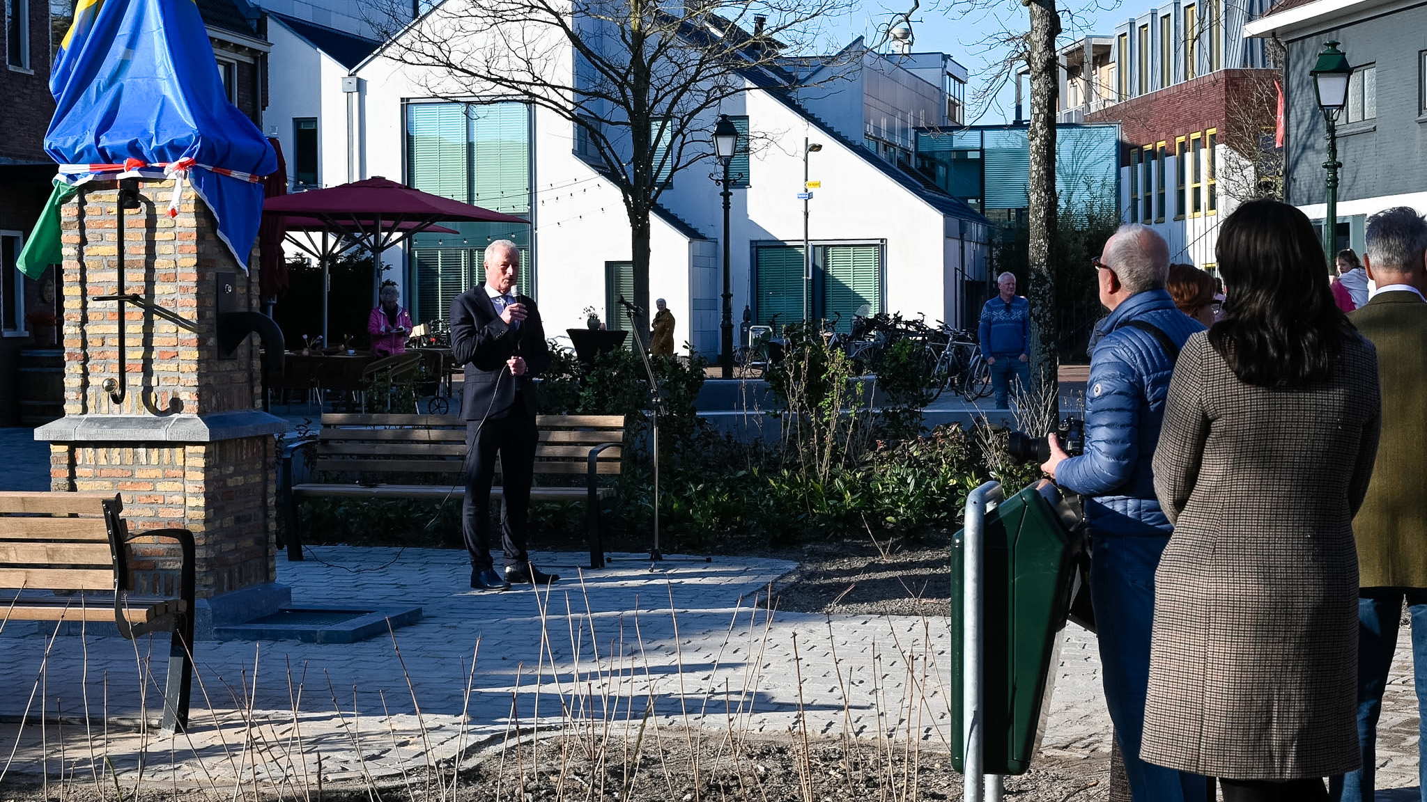 Een man in pak houdt een toespraak naast een bedekt standbeeld, terwijl mensen luisteren op een zonnig plein.