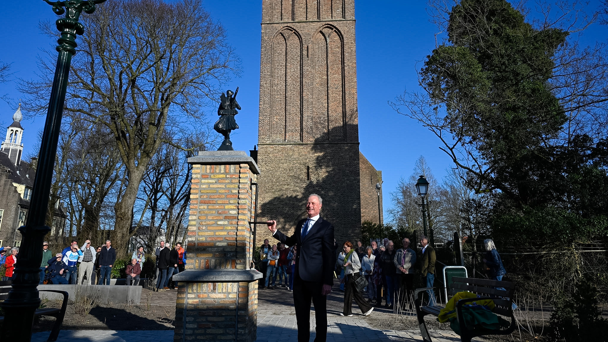 Man onthult een standbeeld op een bakstenen sokkel met een toren op de achtergrond en een groep toeschouwers.