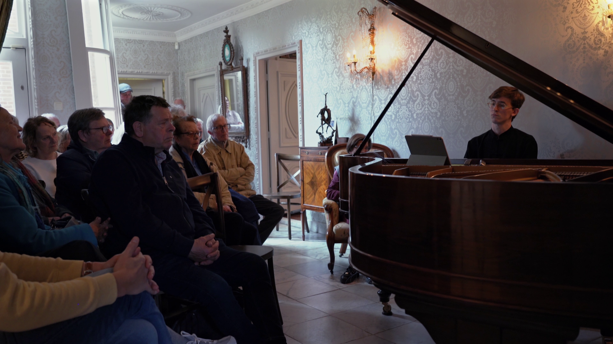Mensen luisteren aandachtig naar een pianist achter een vleugel in een elegant ingerichte kamer.