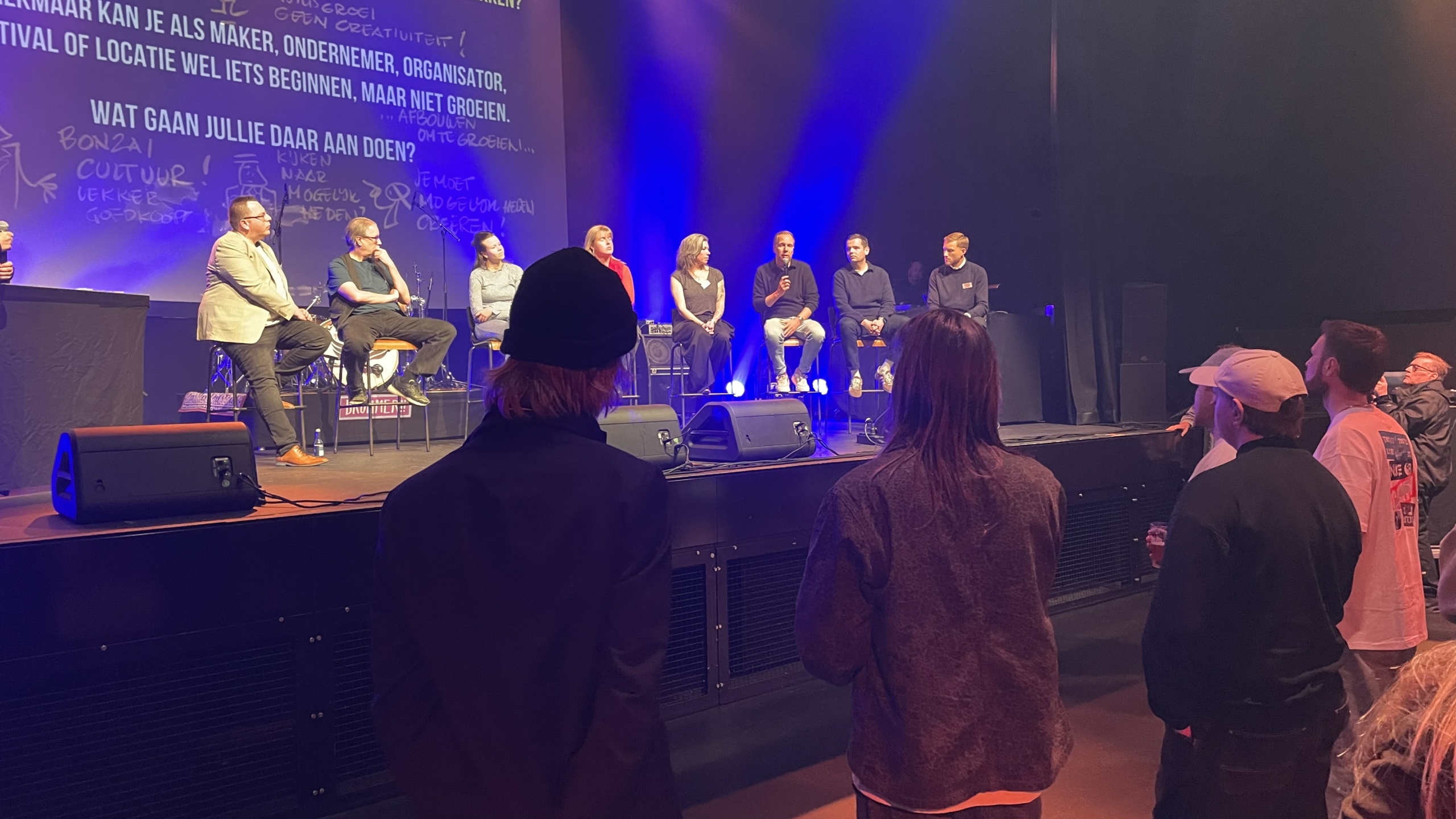 Paneldiscussie op een podium met acht mensen zittend op krukken, omringd door een publiek.