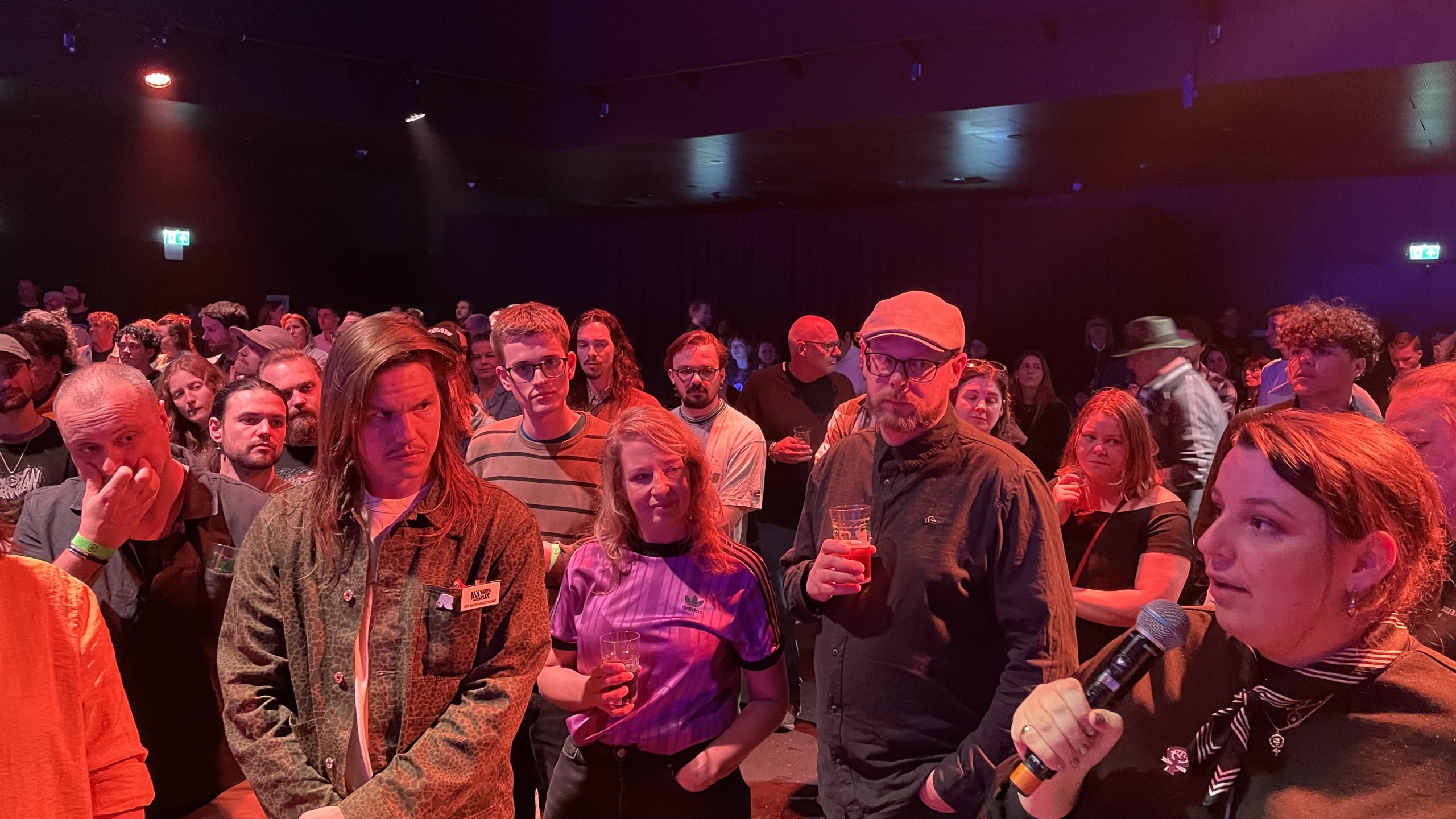 Mensen in een zaal luisteren aandachtig naar iemand met een microfoon; de verlichting is warm en rood.