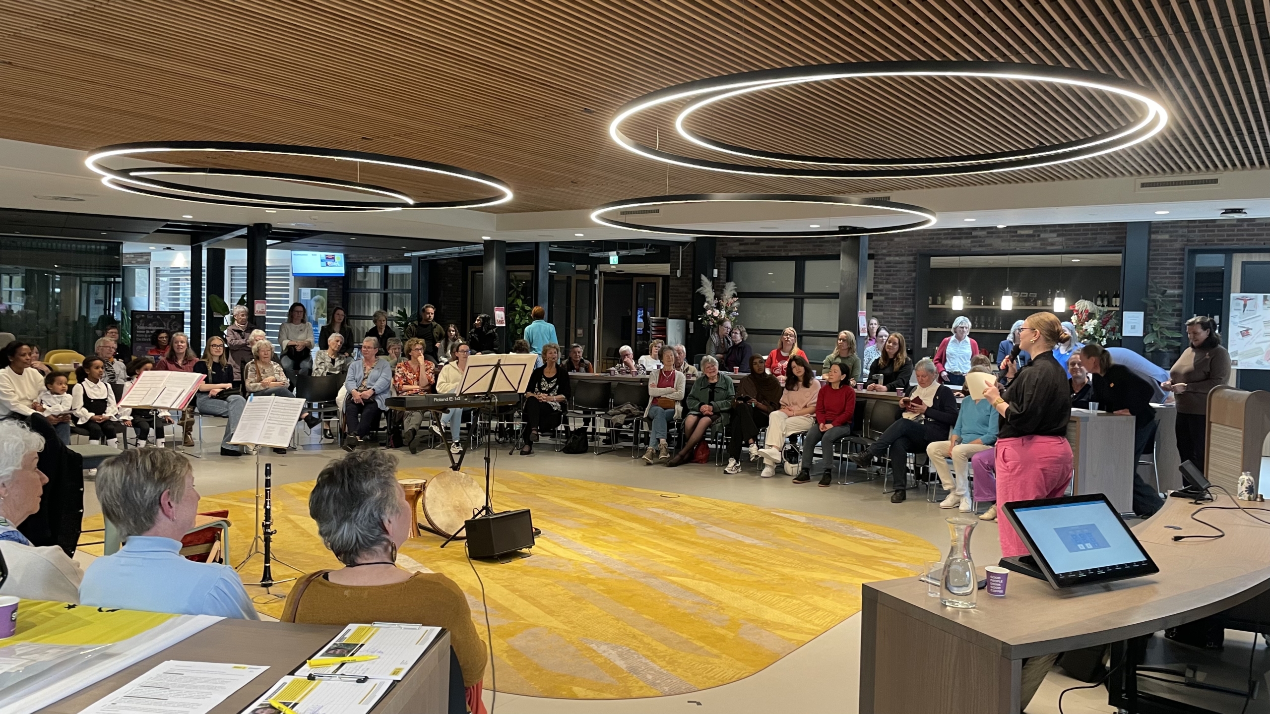 Een grote groep mensen kijkt naar een persoon die spreekt in een moderne ruimte met houten plafond en cirkelvormige lampen.