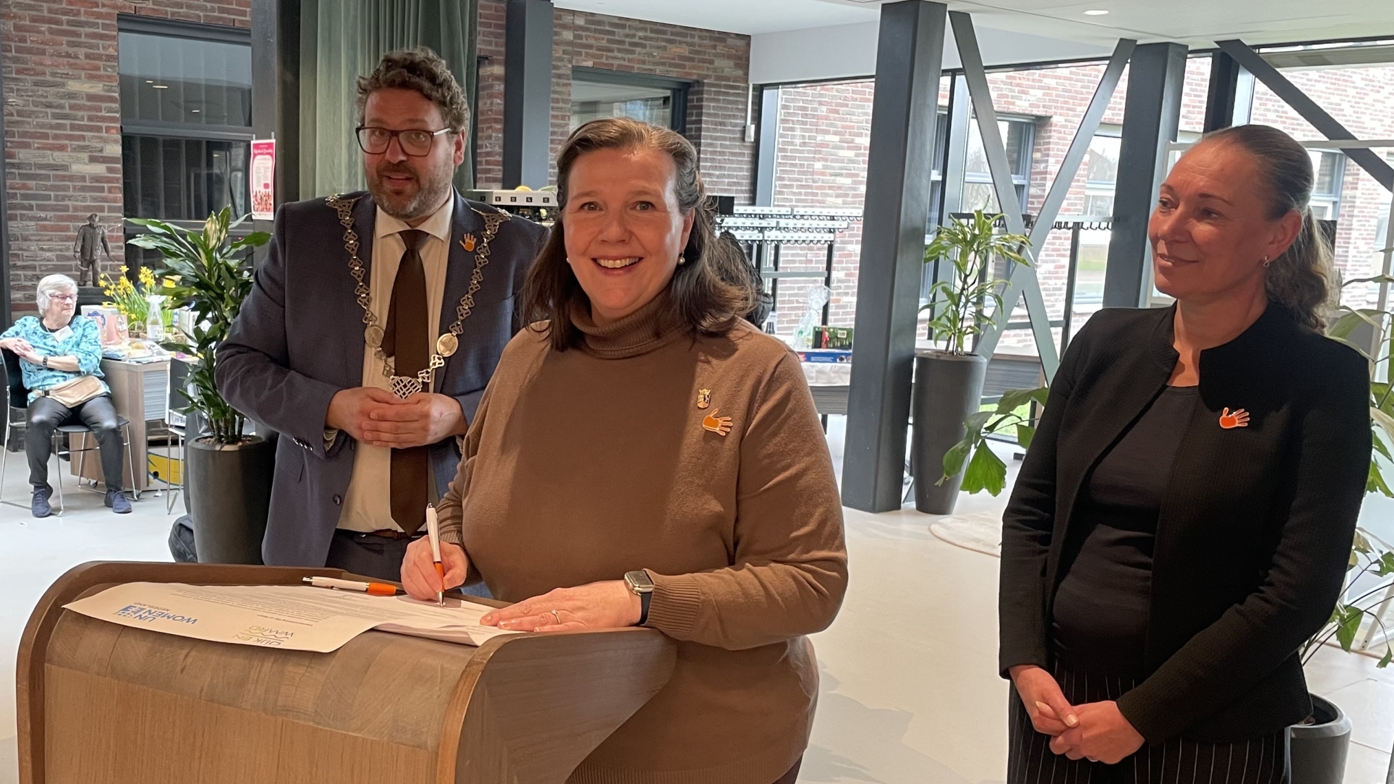 Een vrouw tekent een document op een lessenaar, terwijl anderen om haar heen staan in een moderne ruimte.