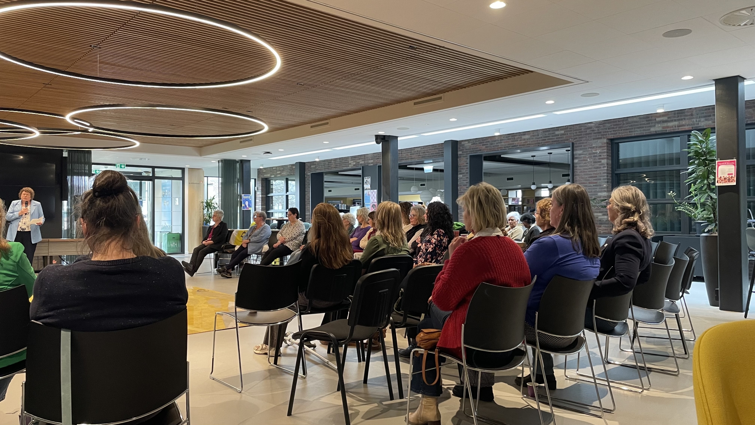 Mensen zitten in een moderne ruimte te luisteren naar een spreker in een lichtblauw jasje.