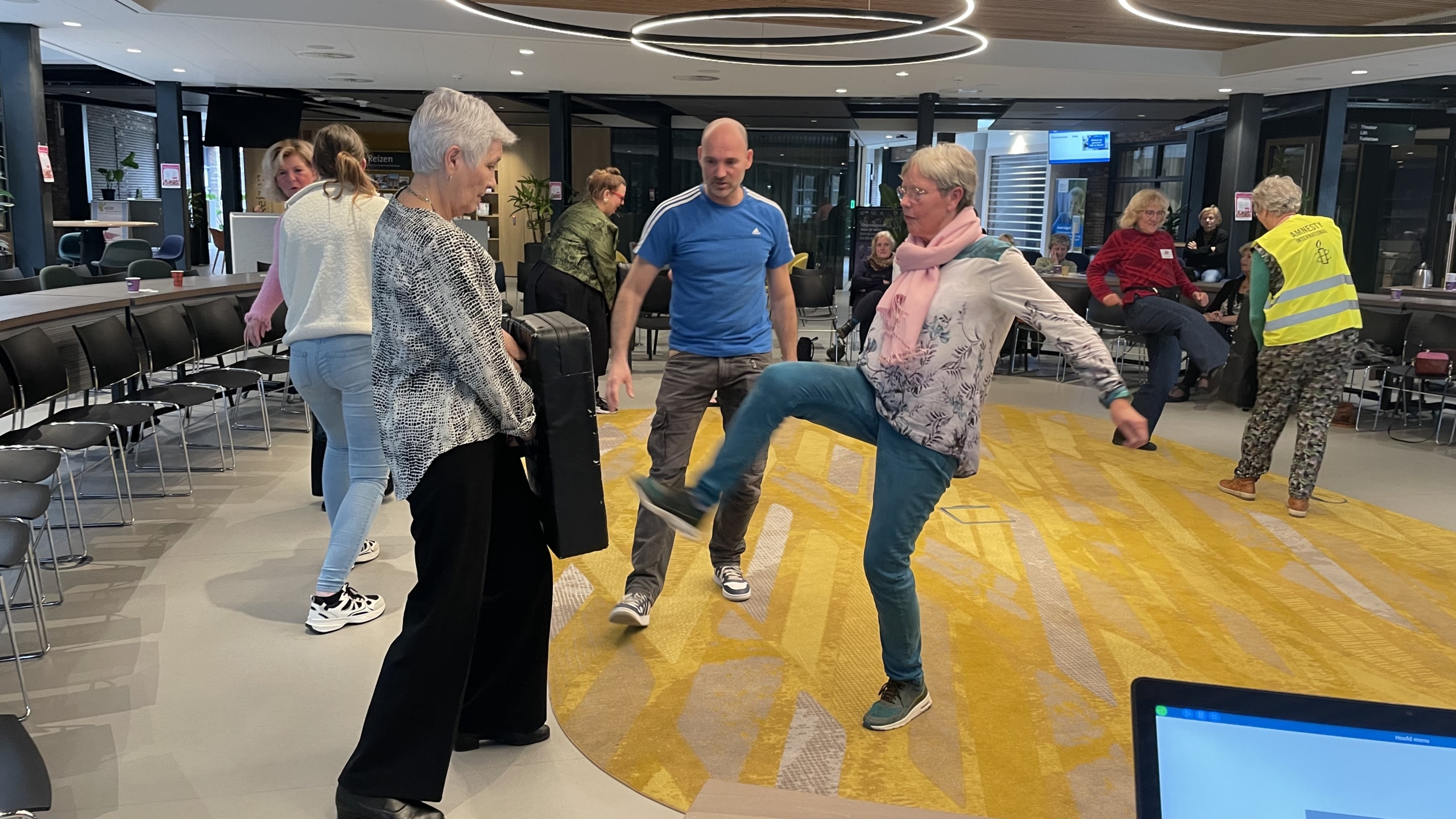Mensen bewegen en dansen in een kring in een open ruimte met stoelen en tafels, met een opvallend geel tapijt.