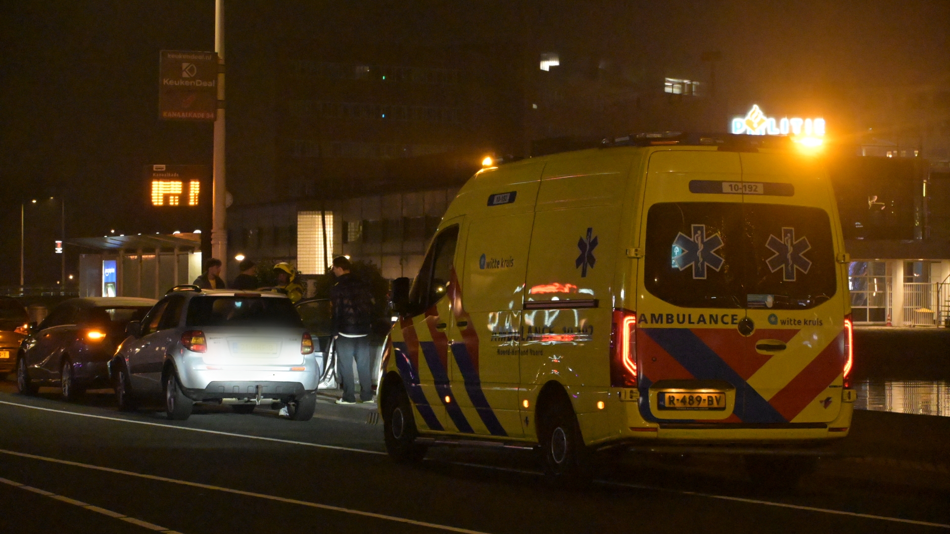 Ambulance en personenauto's langs de weg 's nachts met mensen erbij, in een verlichte stedelijke omgeving.