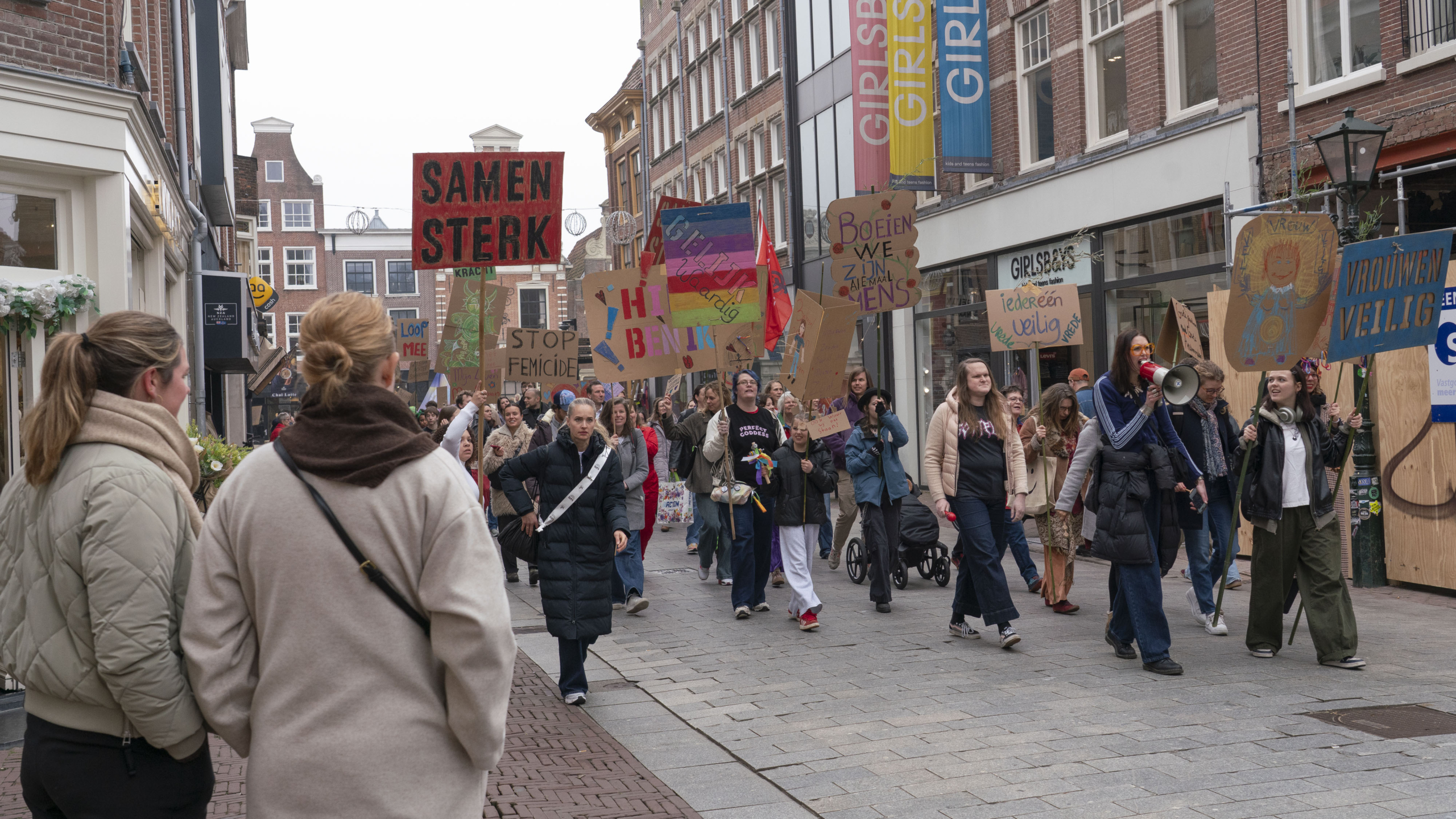 Mensen marcheren in een straat met borden met teksten zoals "Samen Sterk" en "Stop Femicide", die oproepen tot vrouwenrechten.