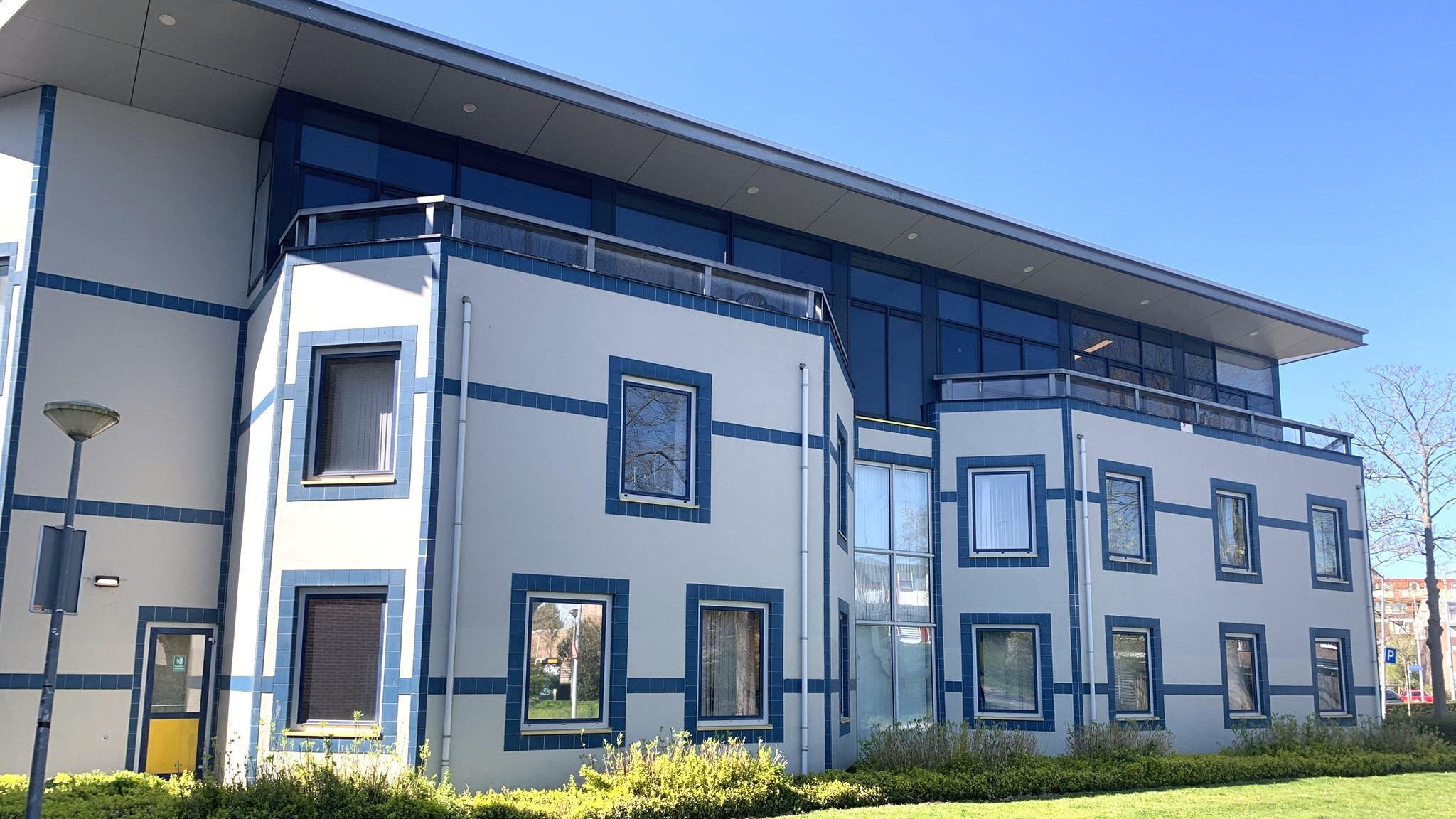 Modern gebouw met blauwe accenten en grote ramen, omgeven door een grasveld en struiken, onder een heldere blauwe lucht.