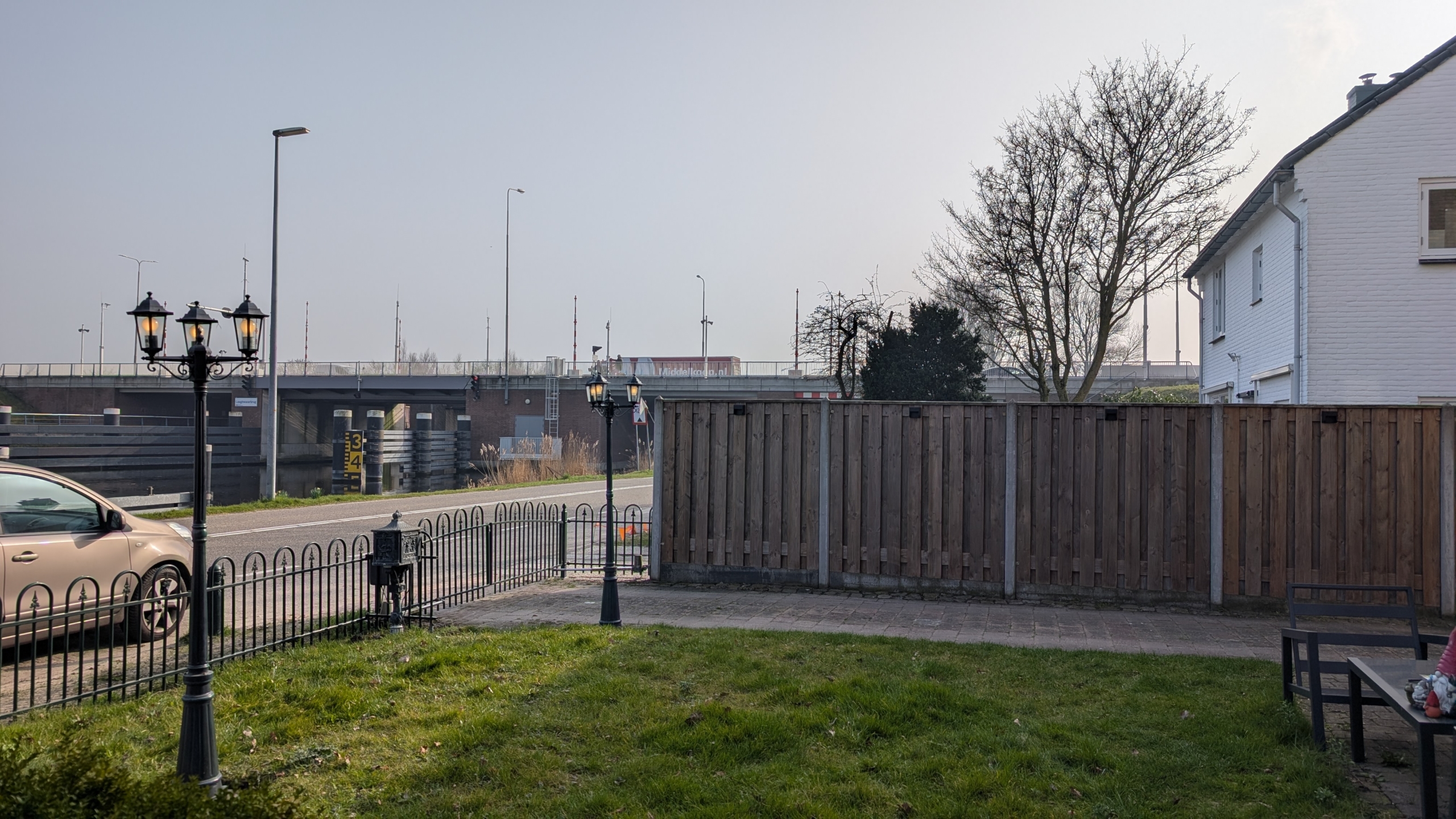 Tuin met een houten schutting, een metalen hek, en twee lantaarns met daarachter een snelweg en voorbijrijdende auto's.