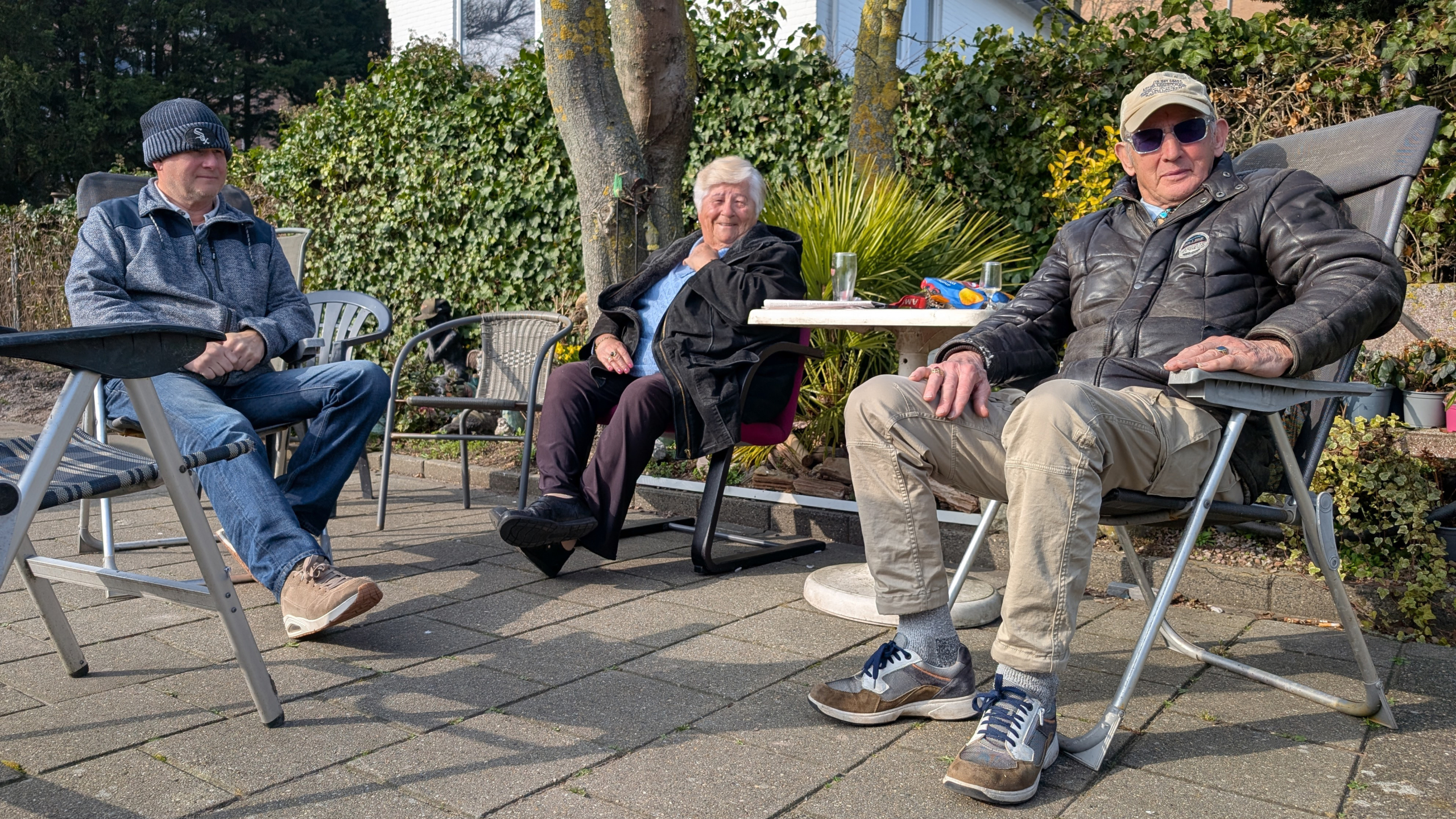 Drie mensen zitten ontspannen in een zonnige tuin, omringd door stoelen en planten.