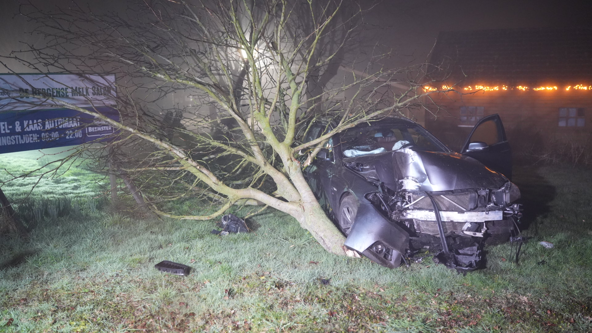 Auto gebotst tegen een boom naast een reclamebord; de voorzijde is zwaar beschadigd, deuren staan open, het is nacht.