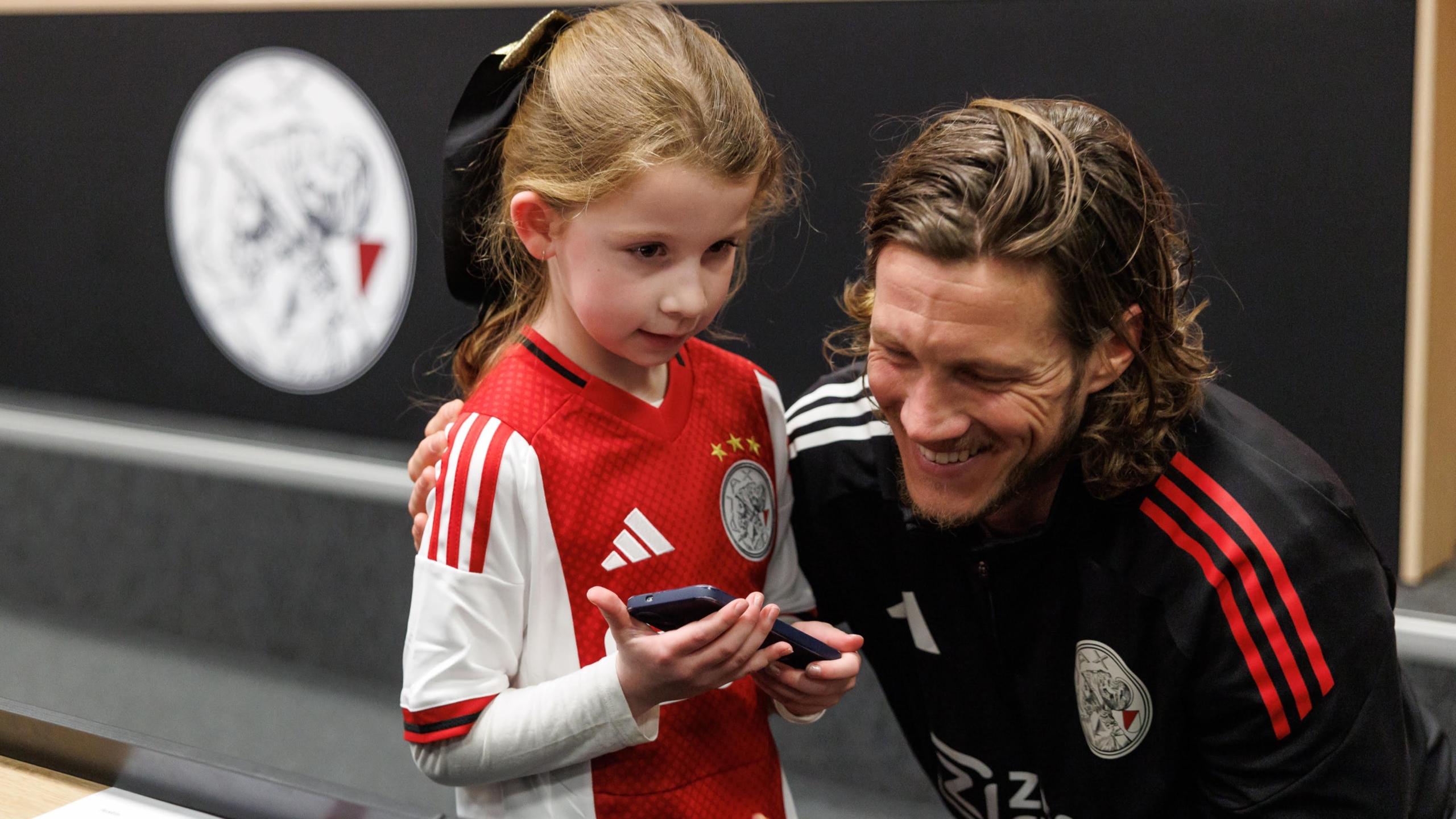 Een meisje in een voetbalshirt met iemand naast haar die haar omarmt en samen naar een telefoon kijken in een sportieve omgeving.