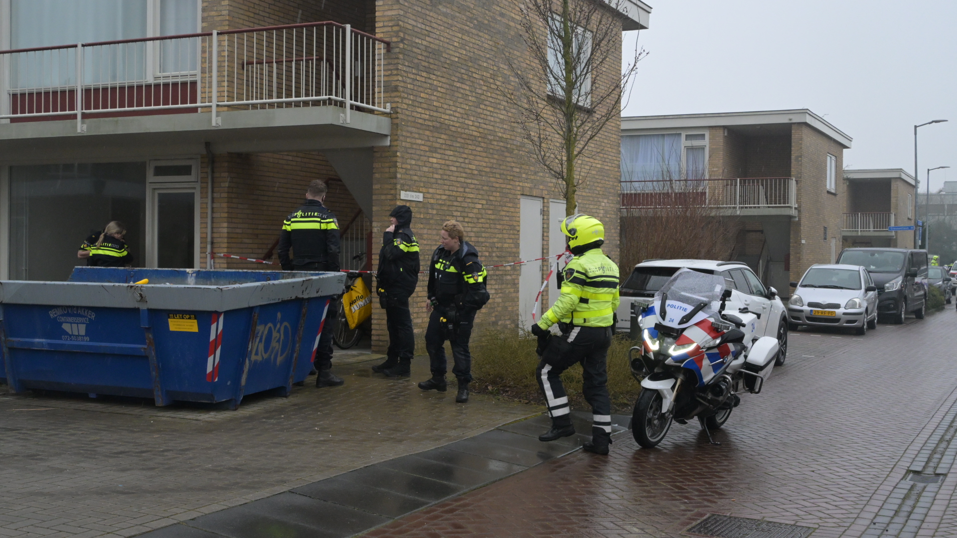 Politieagenten in uniform onderzoeken een plaats delict bij een flatgebouw, met een motor en een container in de buurt.