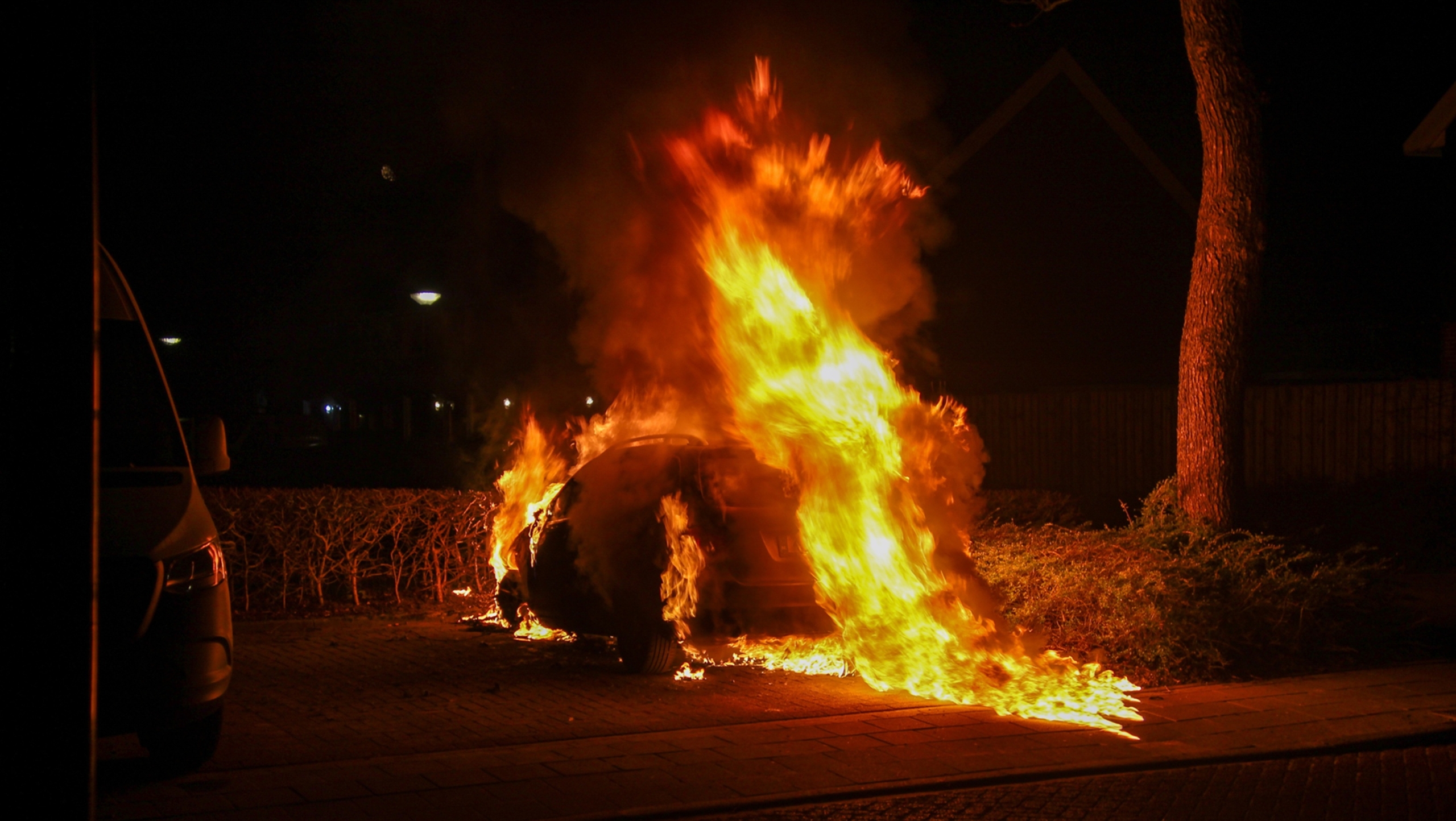 Auto in brand met grote vlammen, naast een boom en een andere geparkeerde auto in een nachtelijke omgeving.