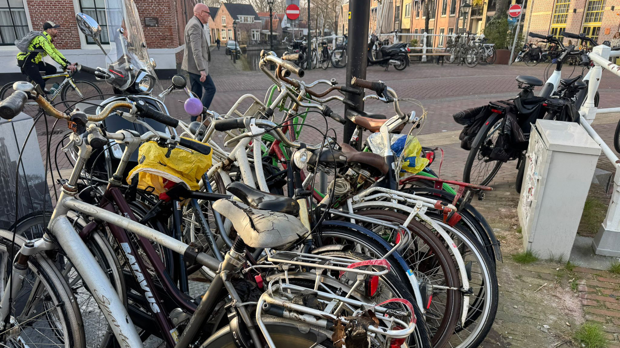 Een groep oude fietsen, waaronder een met een gescheurde zadel, geparkeerd op een steenachtige straat; mensen en andere fietsen op de achtergrond.