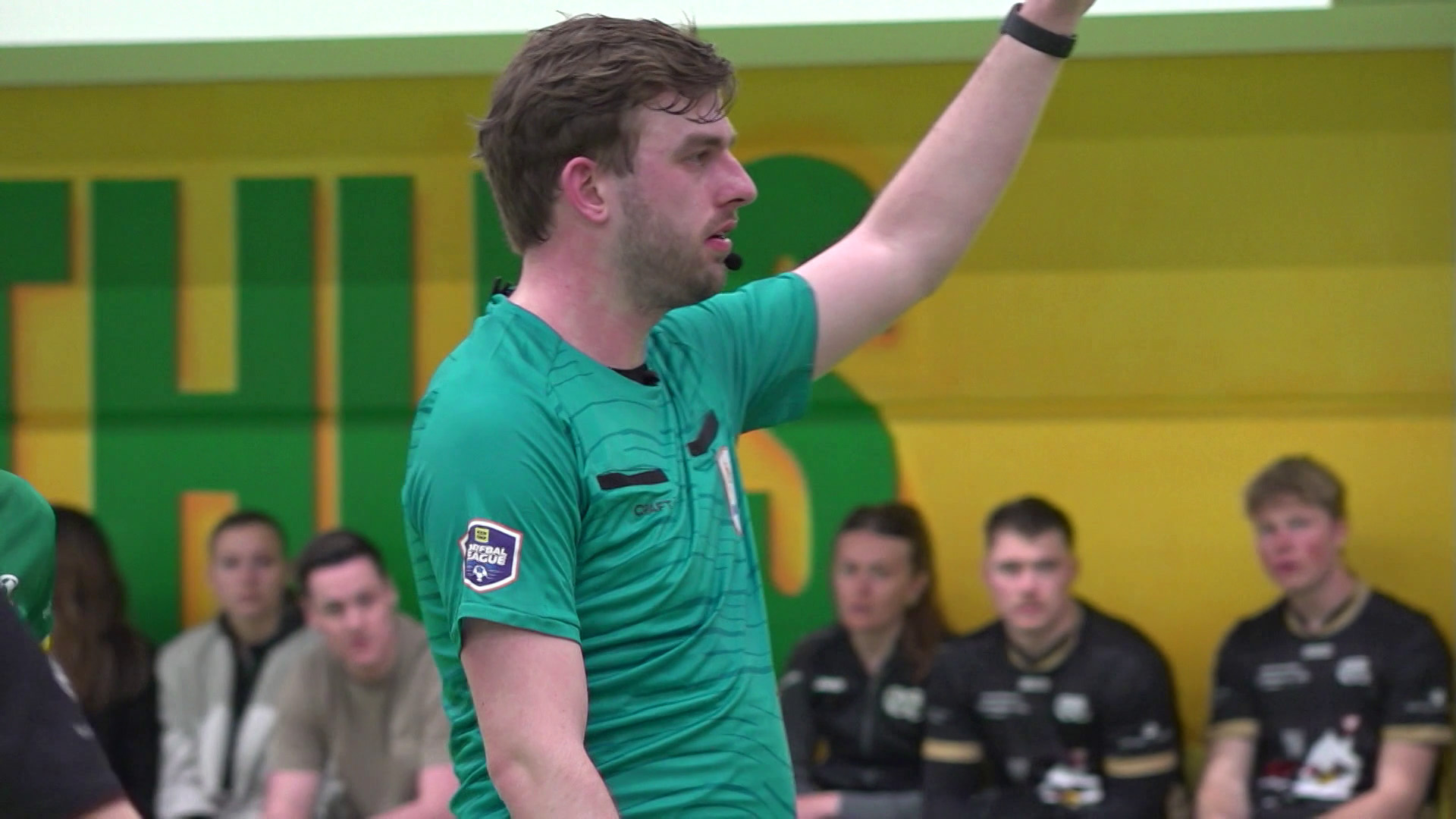 Een scheidsrechter in een groene outfit wijst iets aan, terwijl mensen in sportkleding op de achtergrond zitten.