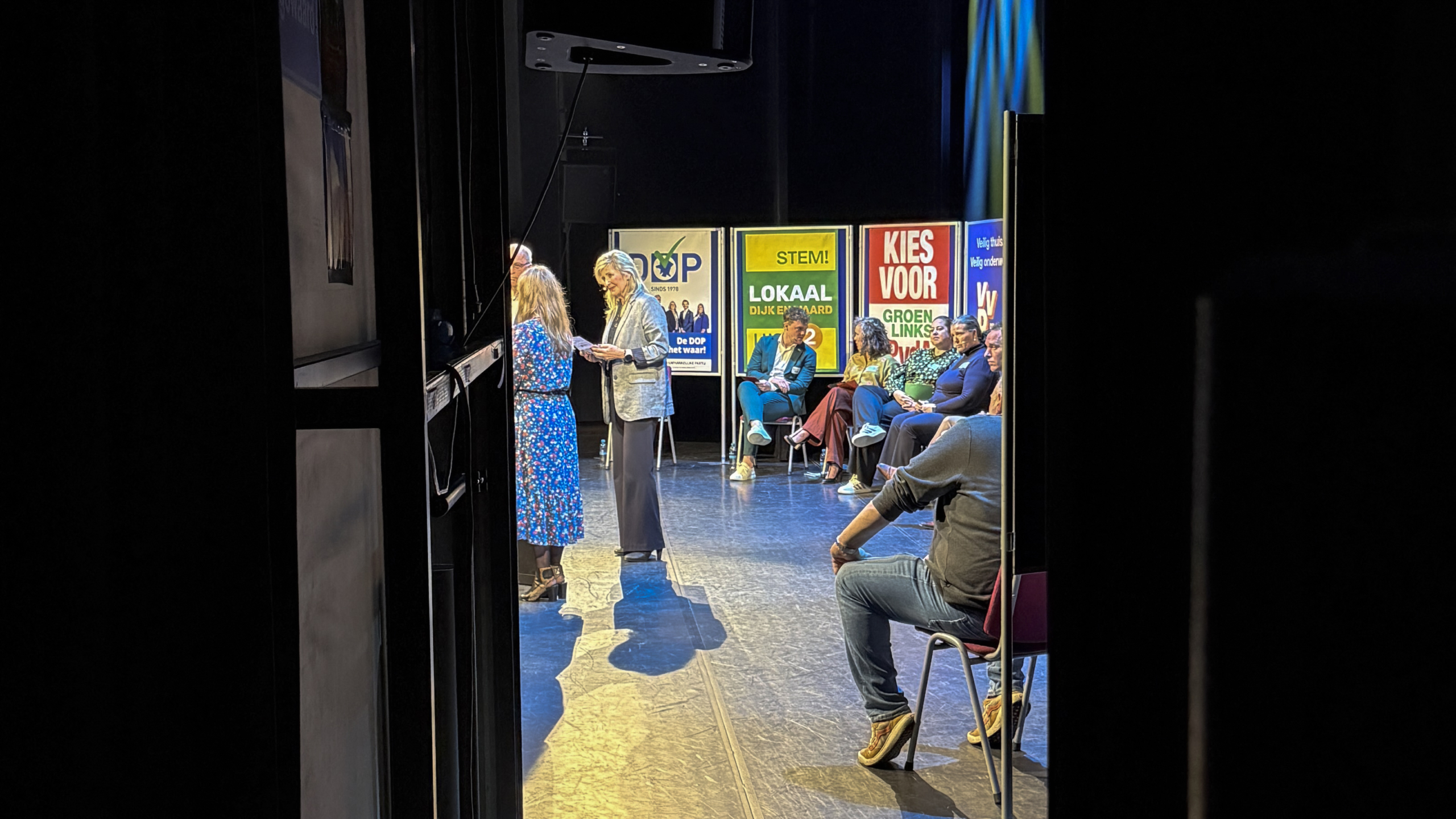 Mensen zitten op een podium tijdens een evenement; op de achtergrond zijn verkiezingsposters zichtbaar.