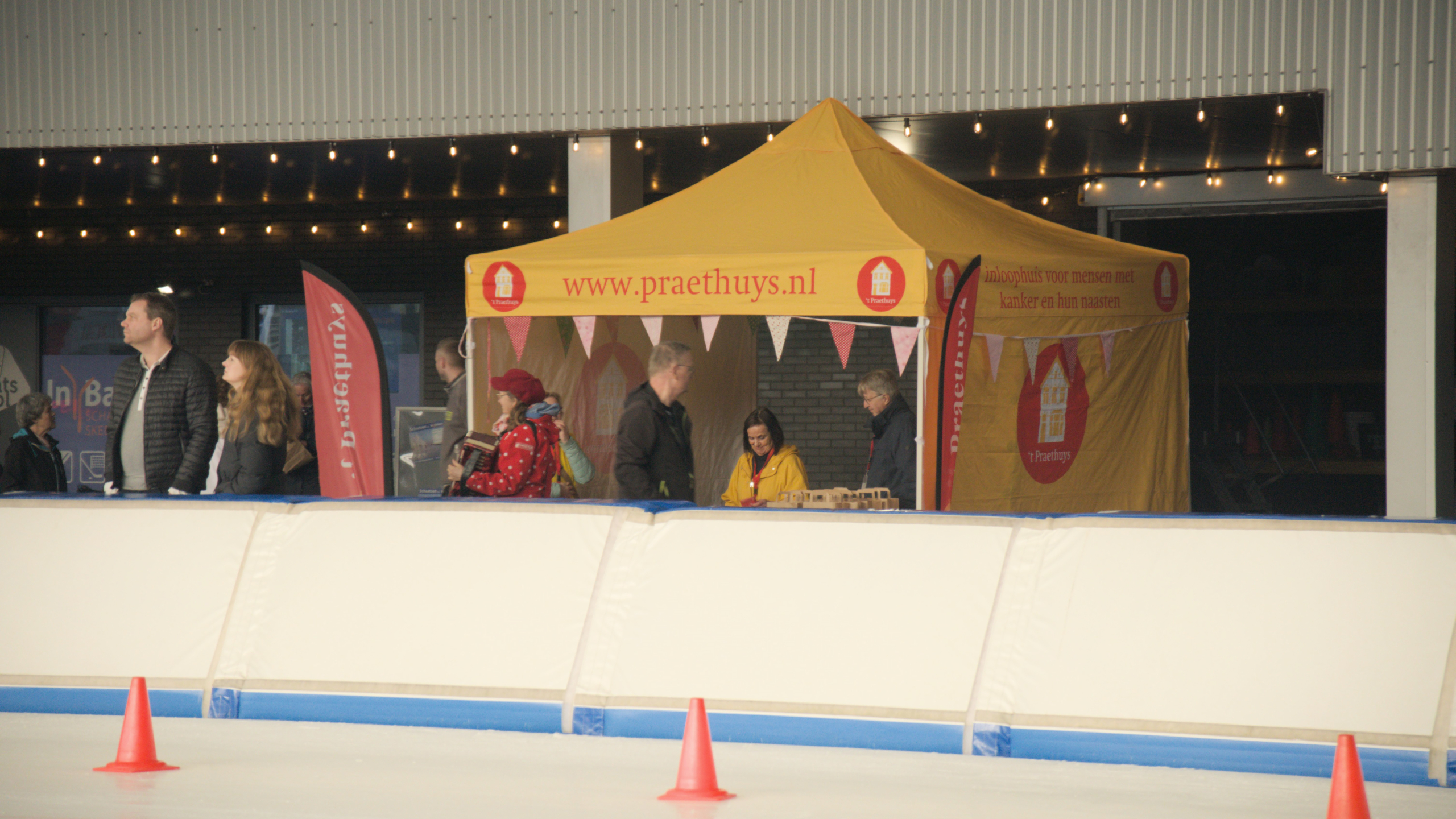 Gele tent met opdruk van Praethuys bij een ijsbaan, omringd door mensen en versieringen, met een oranje pion op de voorgrond.