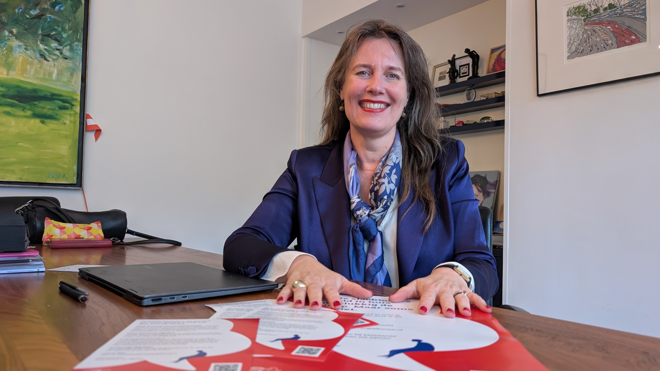 Vrouw zittend aan tafel in een kantoor, lachend naar de camera met documenten en een laptop voor haar.