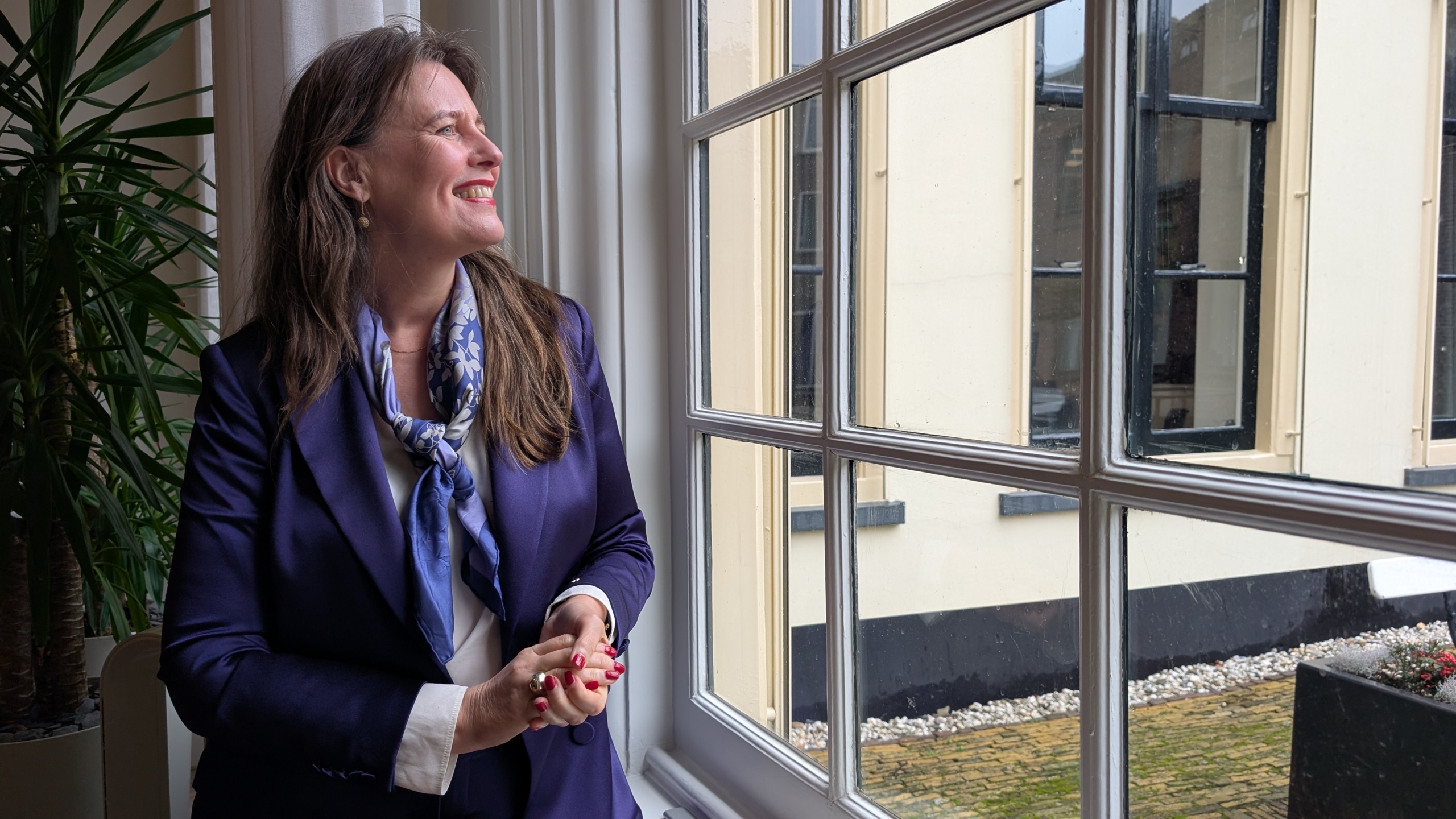 Vrouw in een donkerblauwe blazer kijkt glimlachend uit een raam, met een plant op de achtergrond.