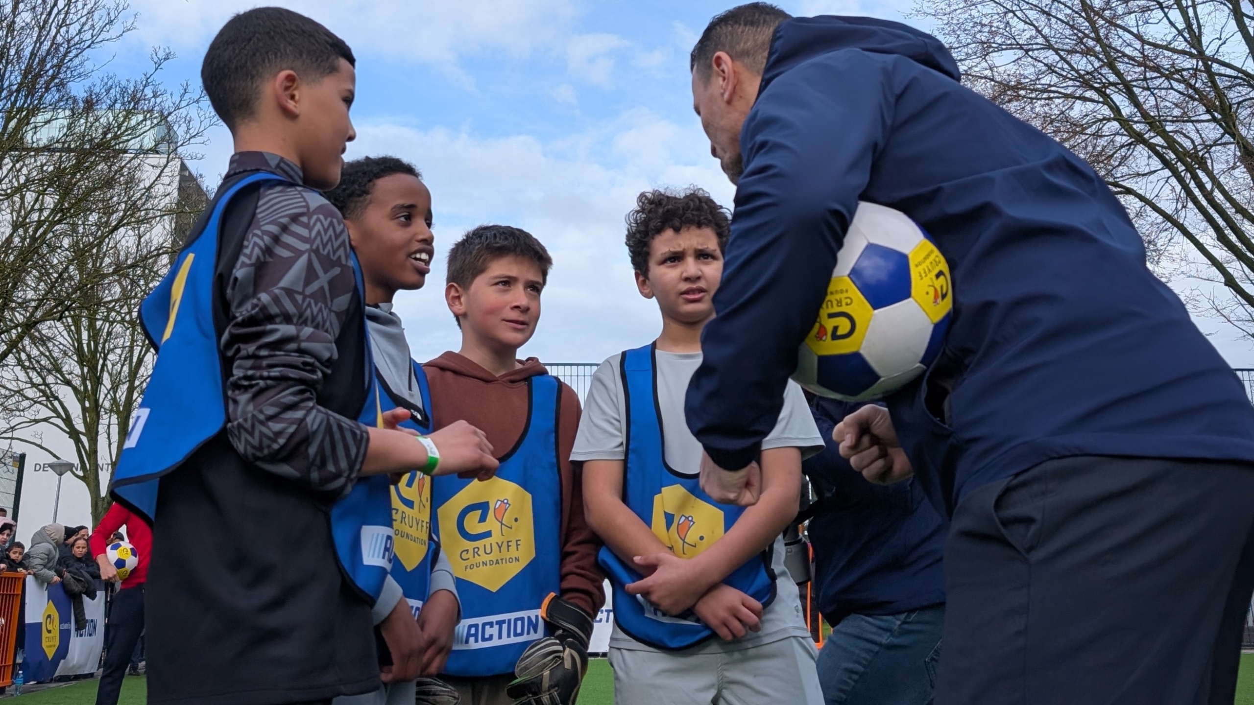 Een groep jongens in blauwe hesjes krijgt instructies van een man met een voetbal op een sportveld.