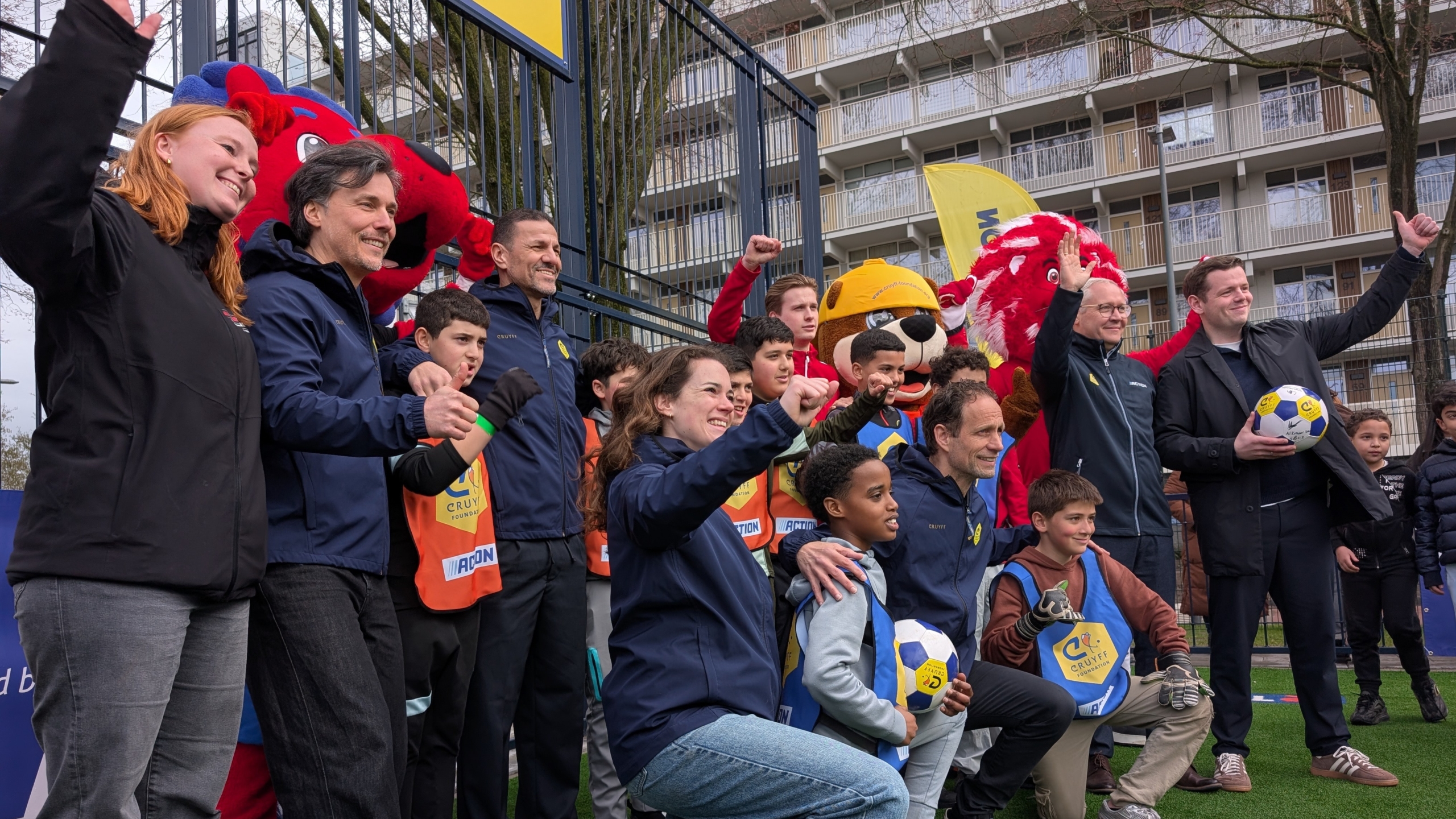 Groep volwassenen en kinderen poseert blij voor de camera op een sportveld, met mascottes en ballen in de omgeving.