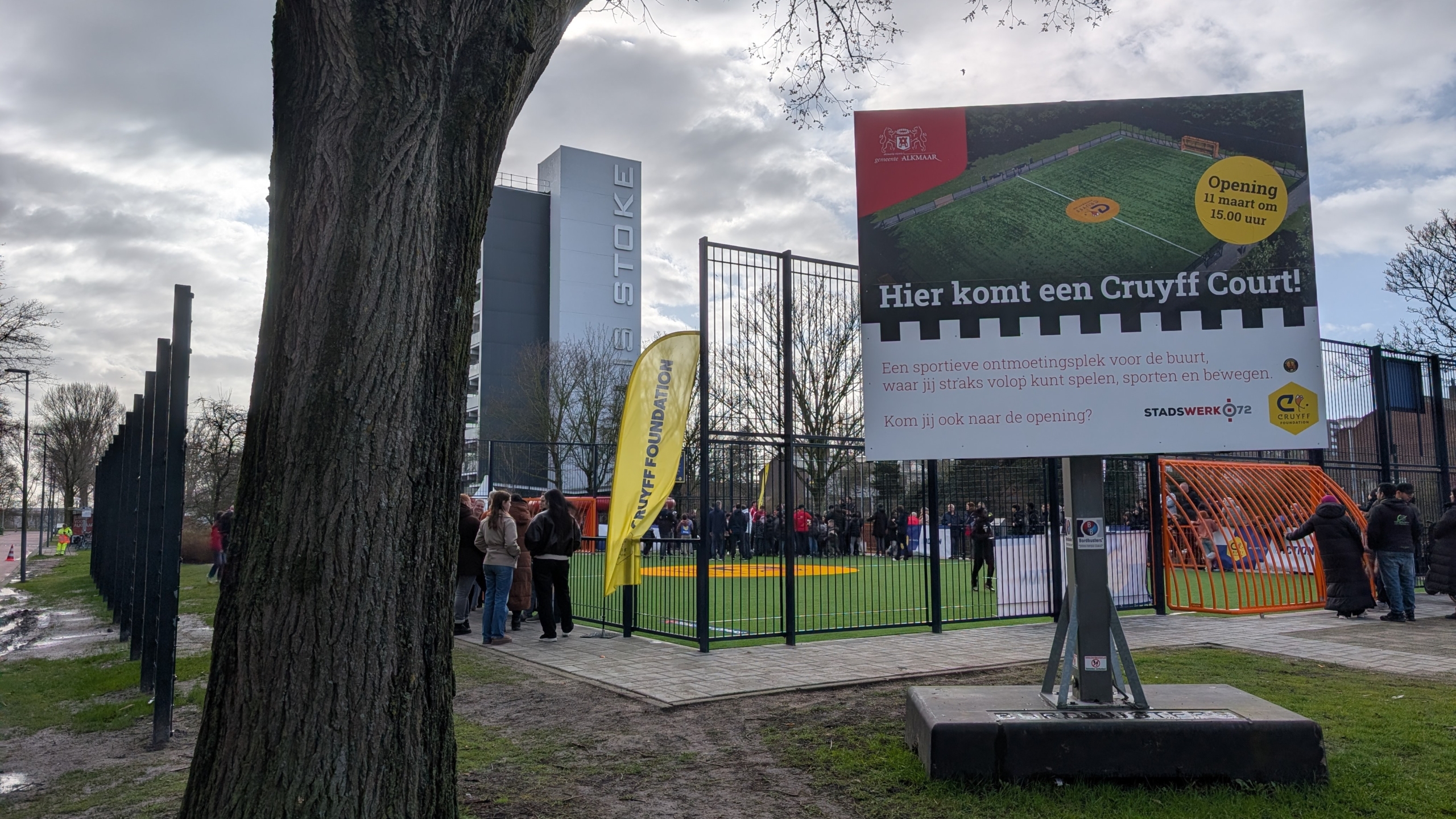 Mensen verzamelen zich bij een Cruyff Court opening; een groot bord en vlag van de Cruyff Foundation zijn zichtbaar.