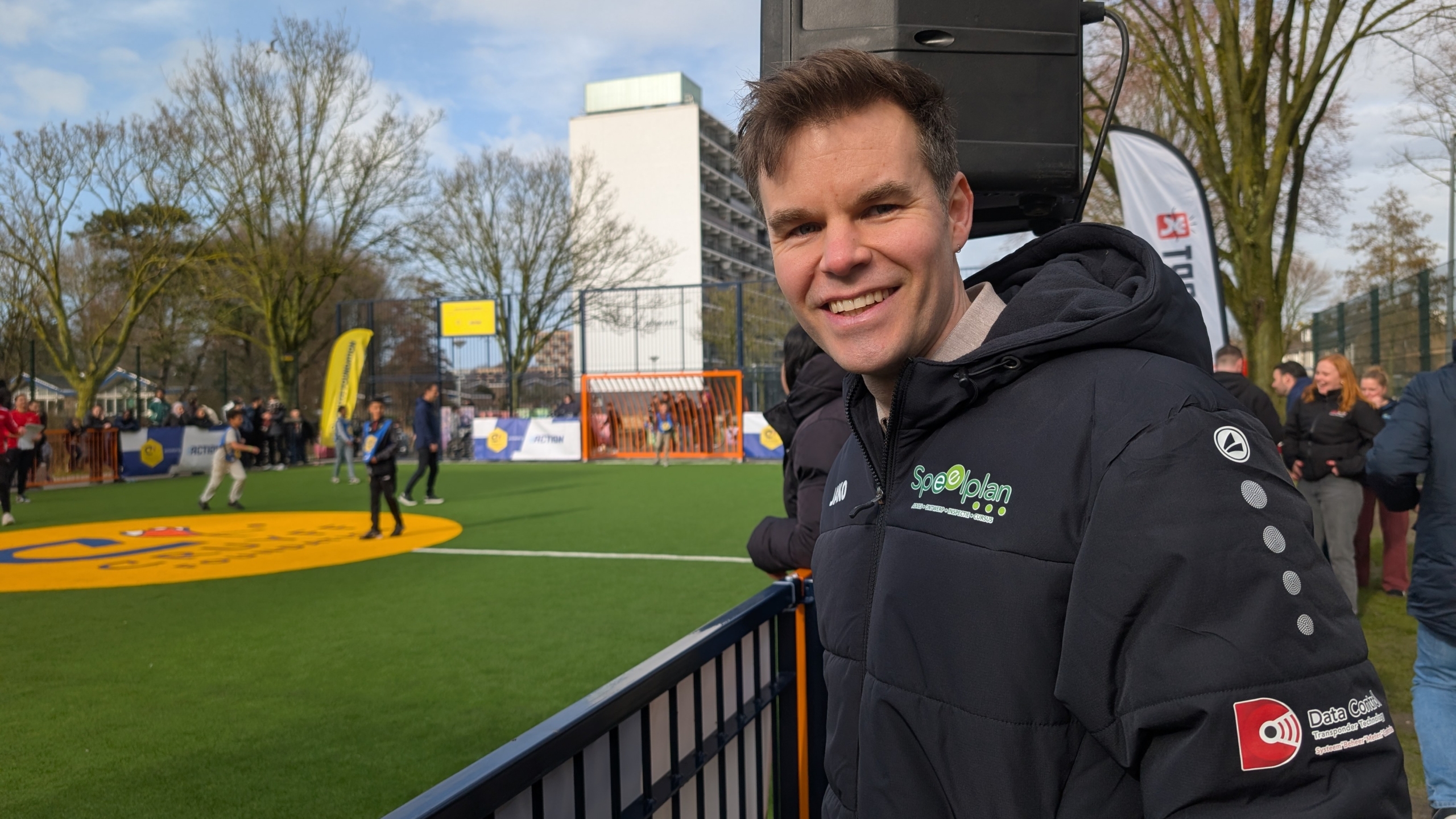 Man lacht op een sportveld met een voetbalwedstrijd op de achtergrond; hij draagt een jas met logo's.