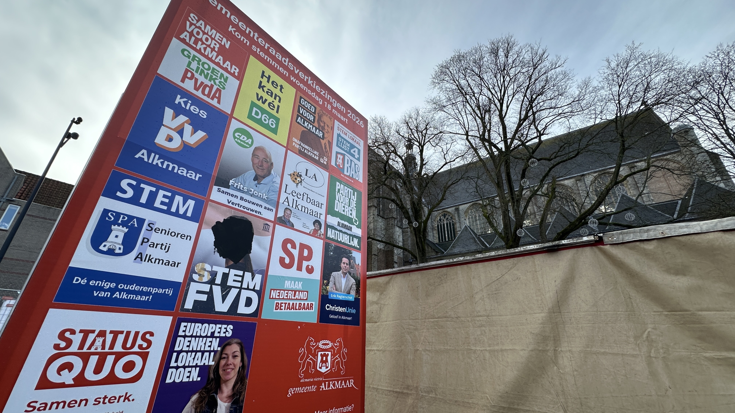 Verkiezingsbord met meerdere partijposters voor de gemeenteraadsverkiezingen 2026 in Alkmaar, tegen de achtergrond van een kerk.