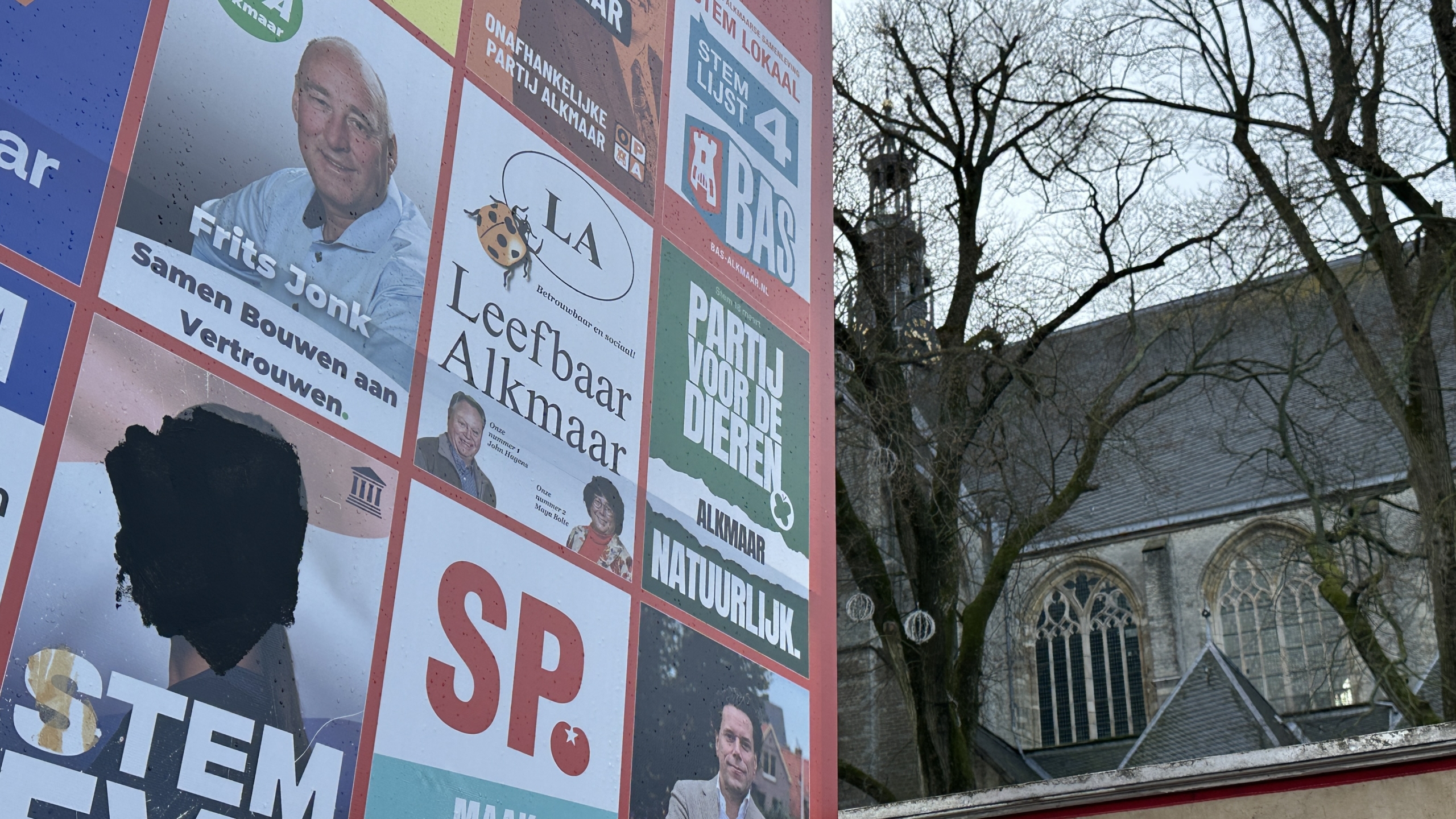 Verkiezingsborden met verschillende politieke partijen en slogans, naast een kerkgebouw onder kale bomen.