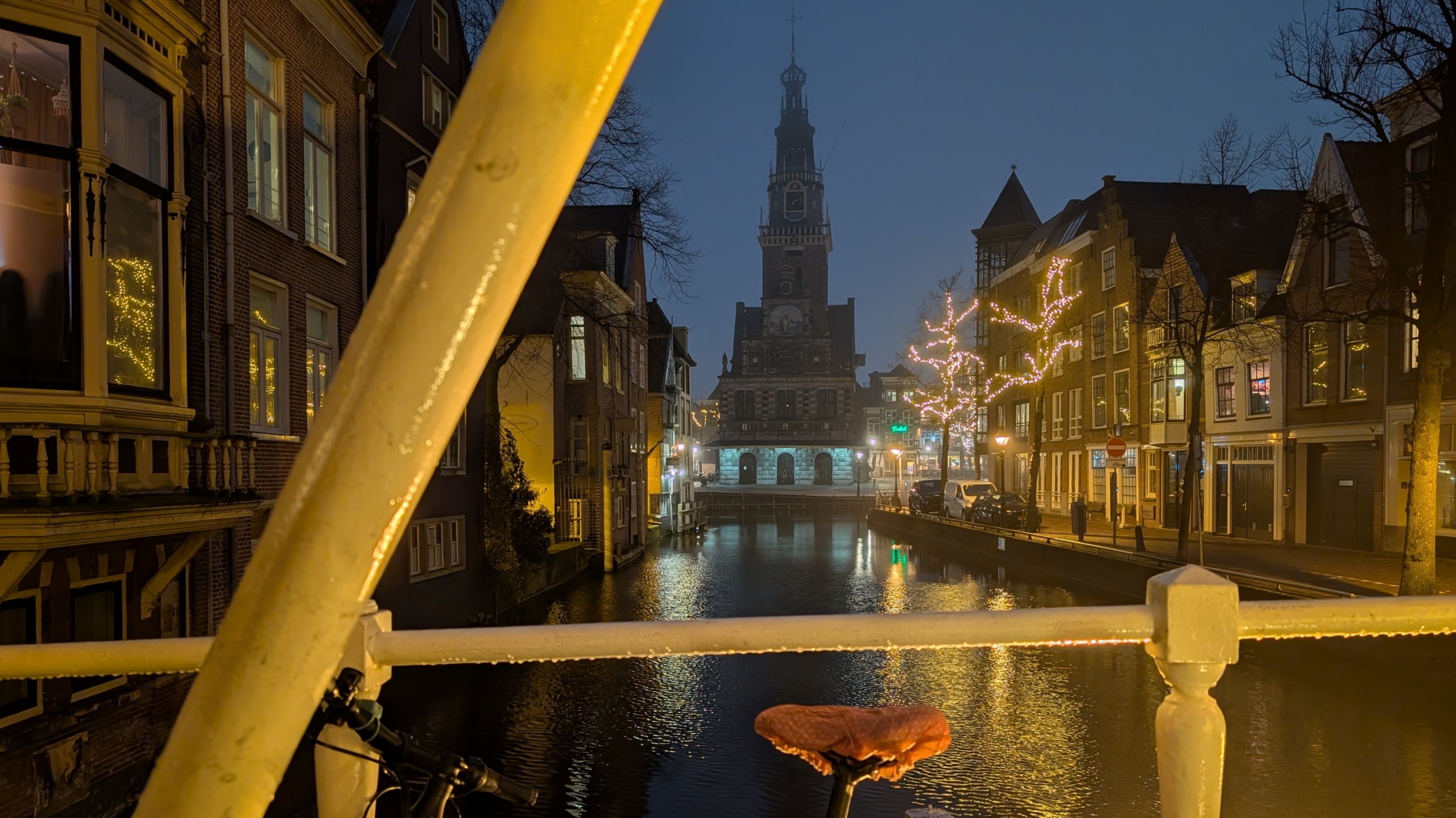 Oude stad in de avond met verlichte gebouwen en een kerk boven een gracht, gezien vanaf een brug met een fiets op de voorgrond.