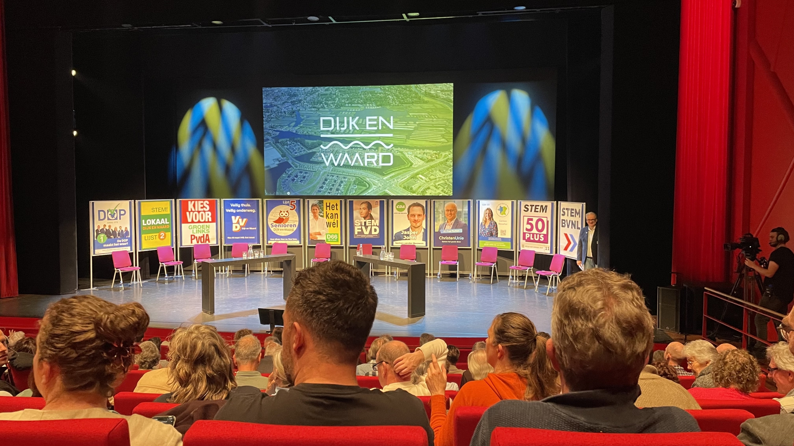Verkiezingsdebat in theater met kleurrijke partijposters, lege stoelen op het podium, publiek en een cameraman.