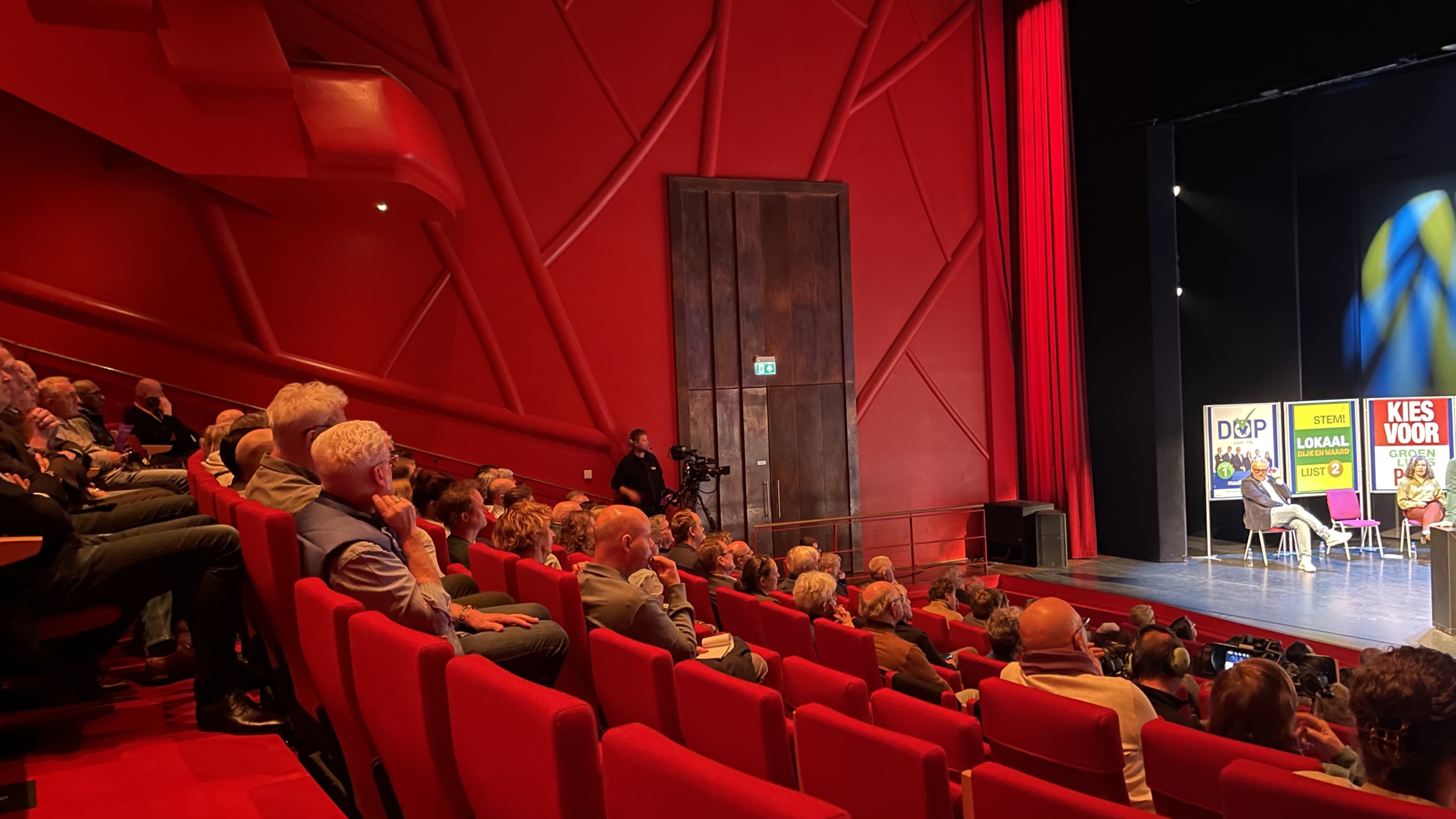 Publiek in een theaterzaal kijkt naar een podium met politieke posters en enkele sprekers.