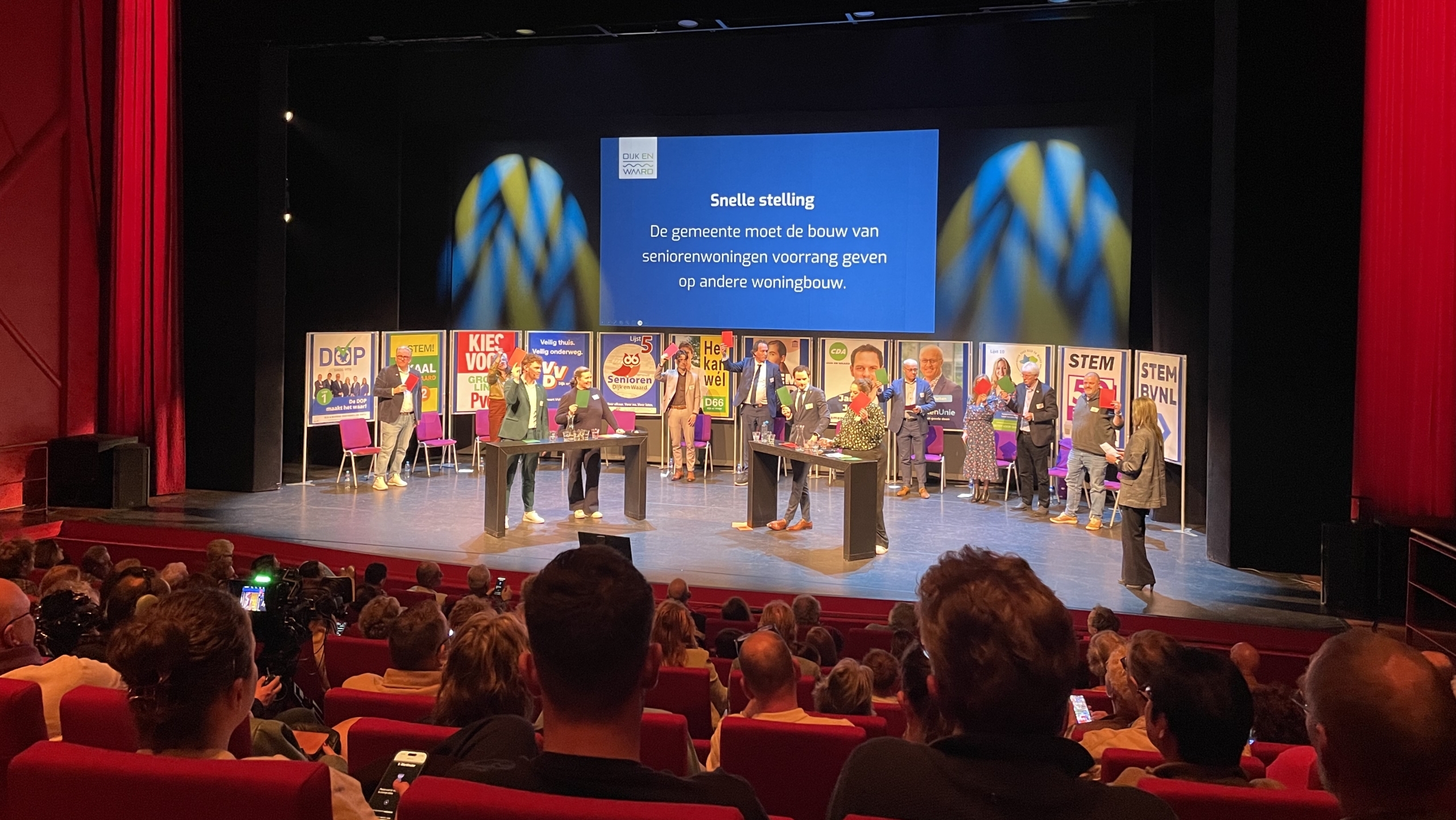 Debat op een podium met meerdere sprekers en een scherm met de tekst over voorrang bij woningbouw. Publiek zit in rode stoelen.