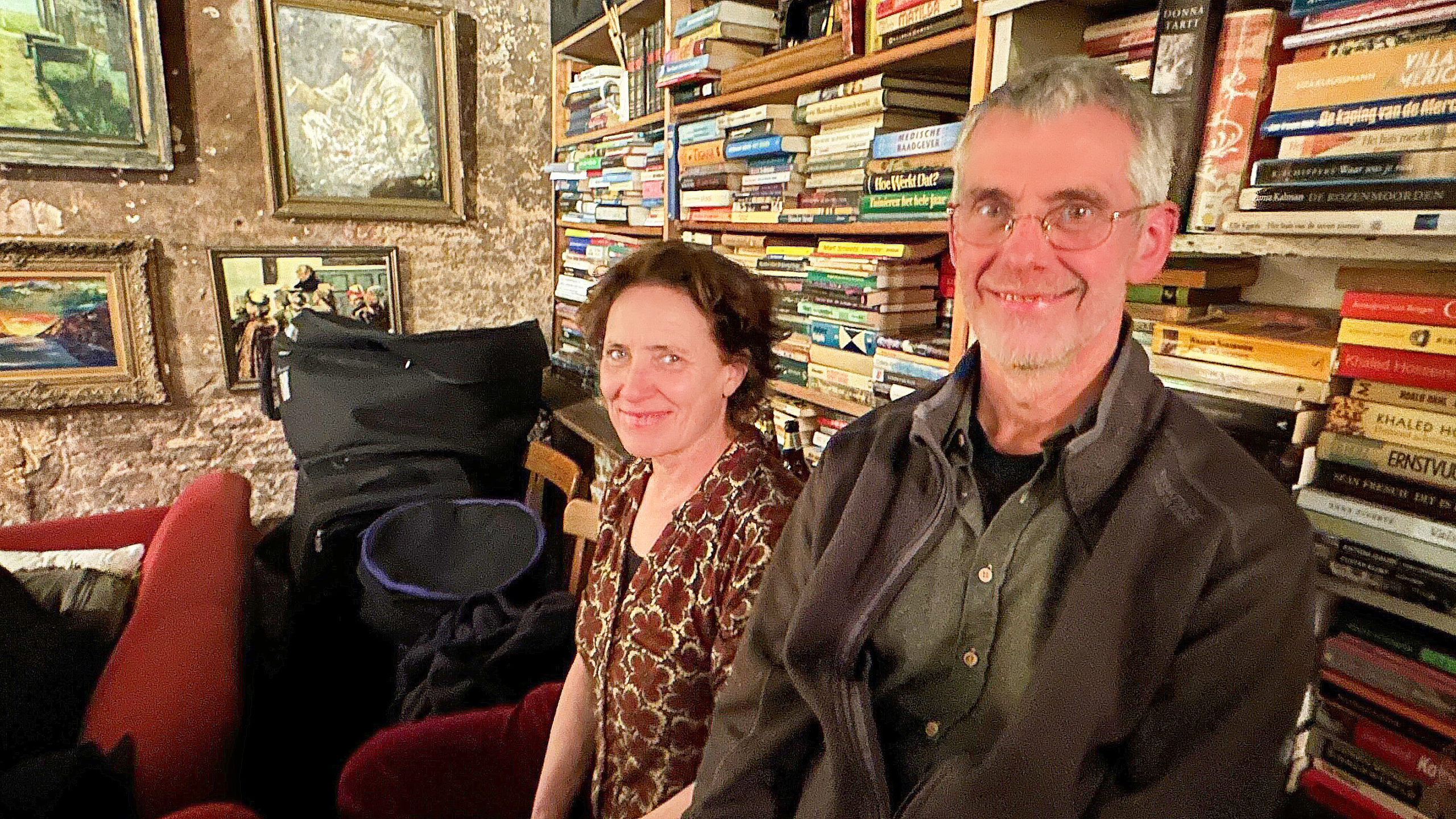 Twee mensen zitten voor een volle boekenkast in een knusse kamer met kunstwerken aan de muur en dragen informele kleding.