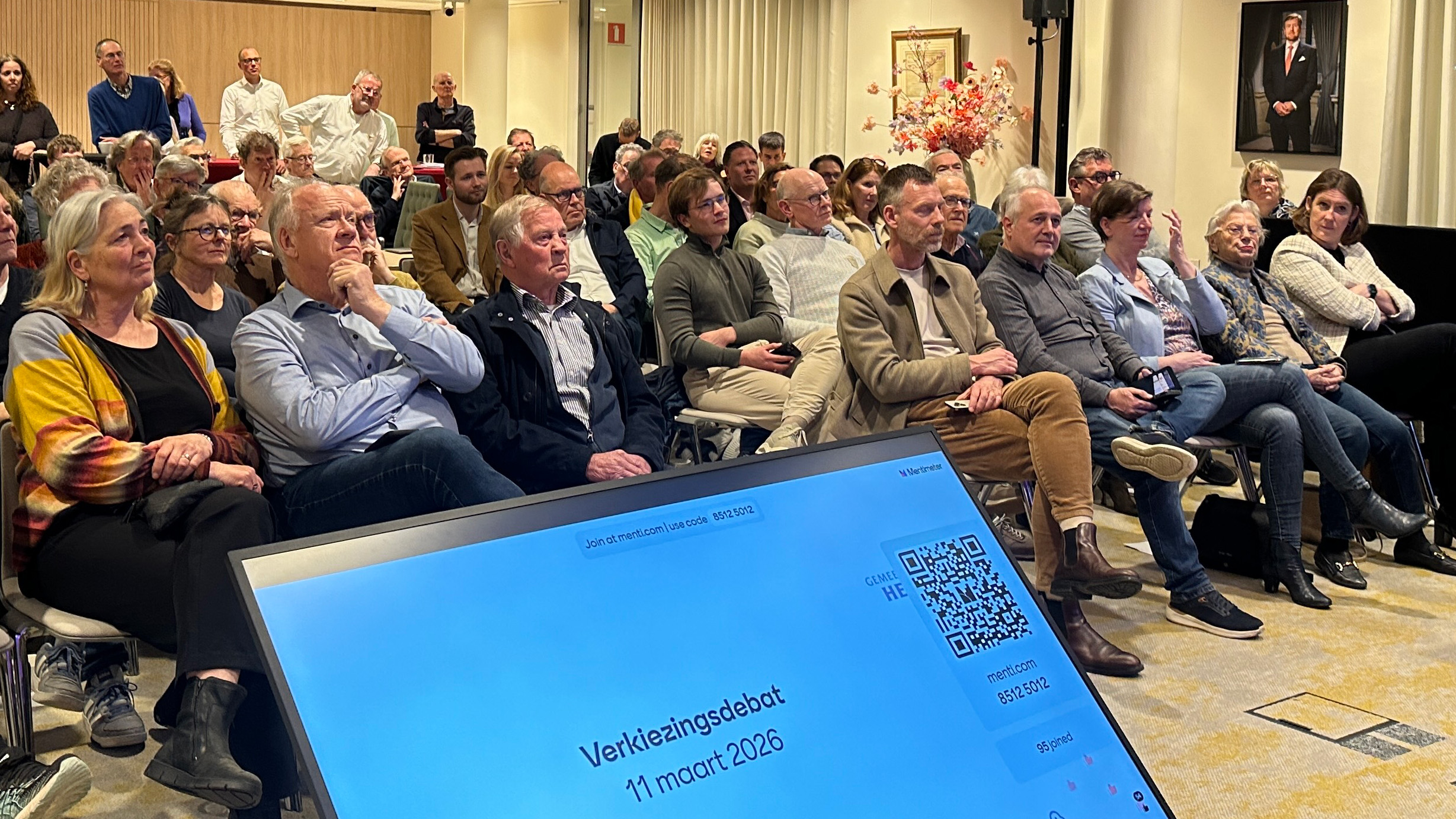 Mensen luisteren naar een verkiezingsdebat op 11 maart 2026; een scherm met details is op de voorgrond zichtbaar.