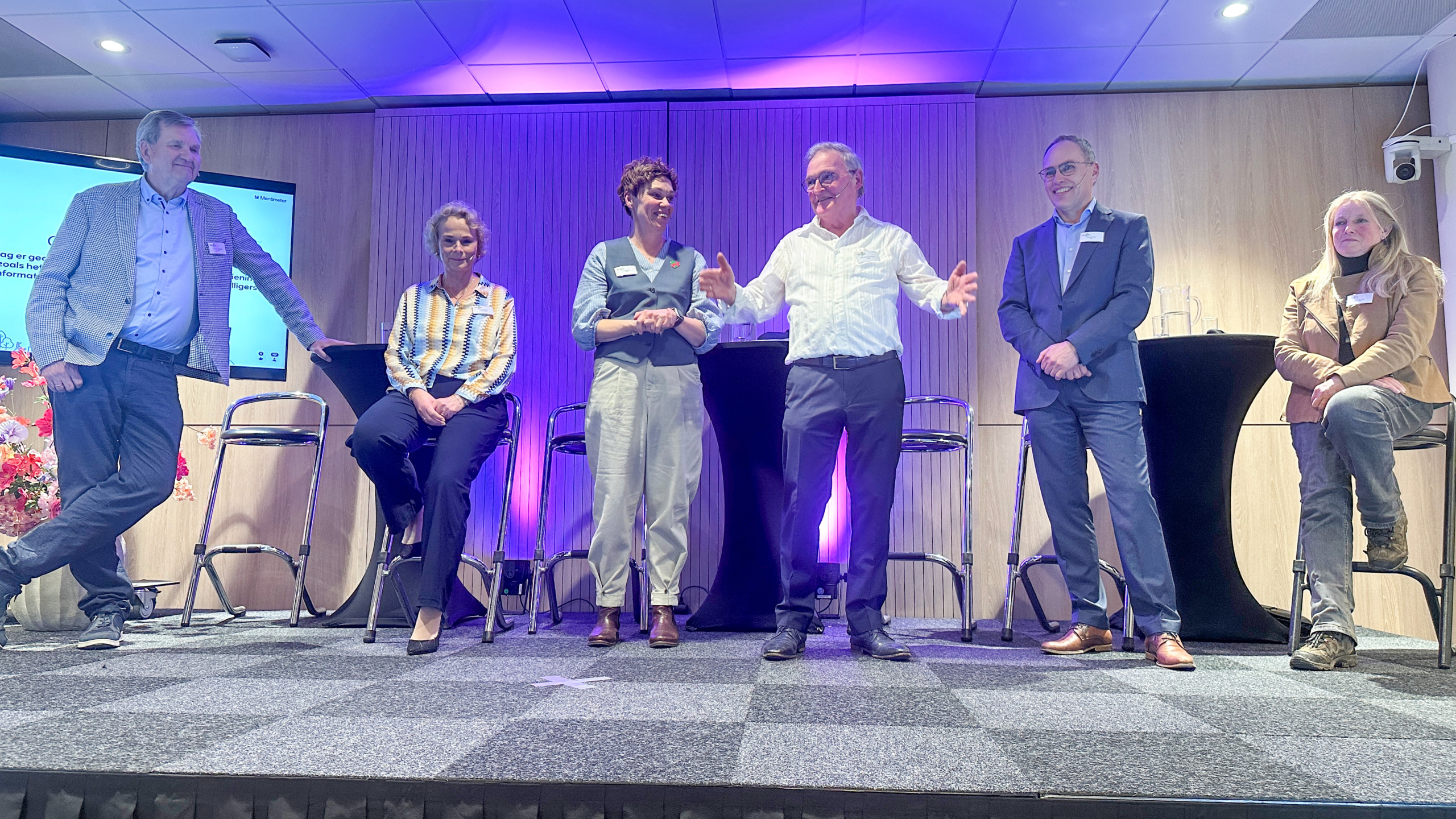 Zes personen staan of zitten op een podium tijdens een conferentie, met een presentatie op de achtergrond en paars licht.