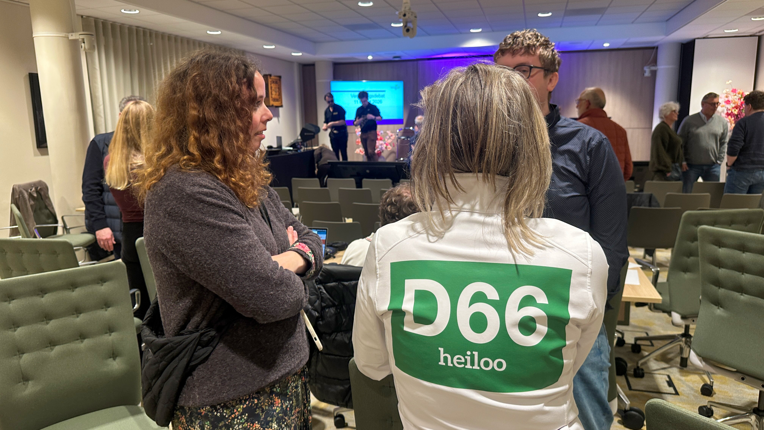 Mensen in gesprek in een zaal na afloop van een evenement; een persoon draagt een shirt met "D66 Heiloo".
