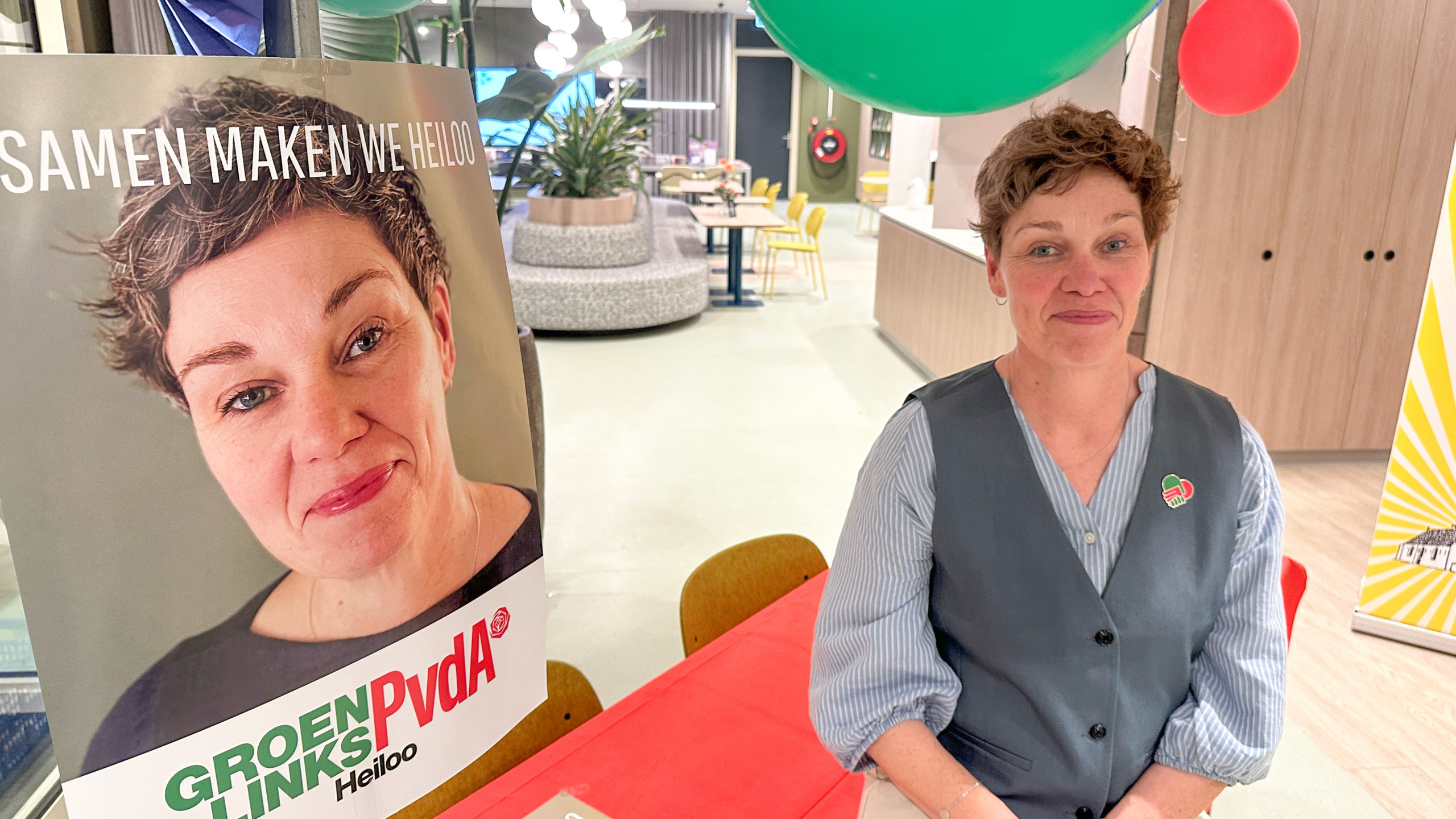 Vrouw zittend naast een verkiezingsposter van GroenLinks PvdA met tekst "Samen maken we Heiloo" in een moderne ruimte.