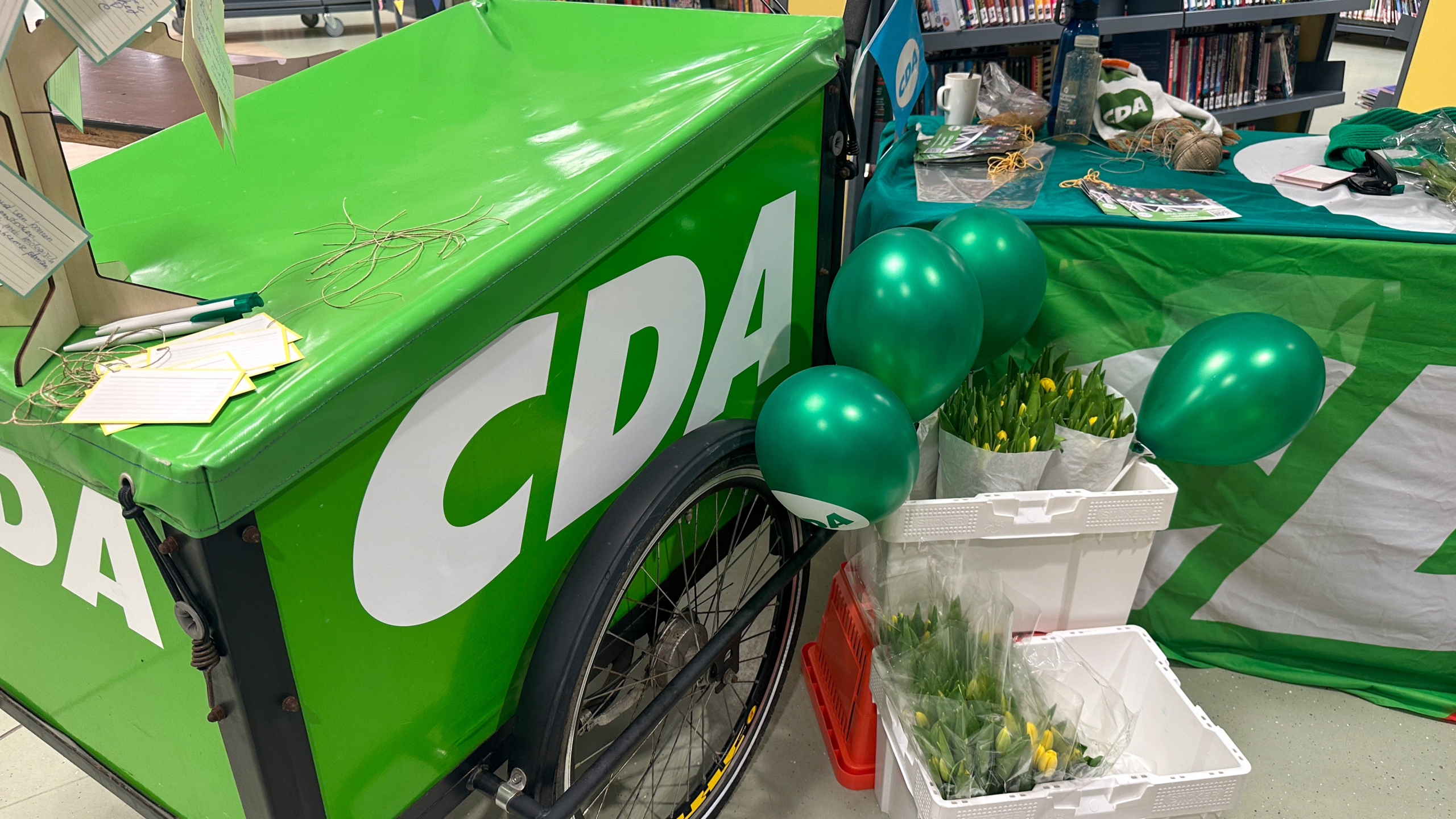 Groene bakfiets met de letters "CDA", omringd door groene ballonnen, tulpen in kratten en promotiemateriaal op tafel.