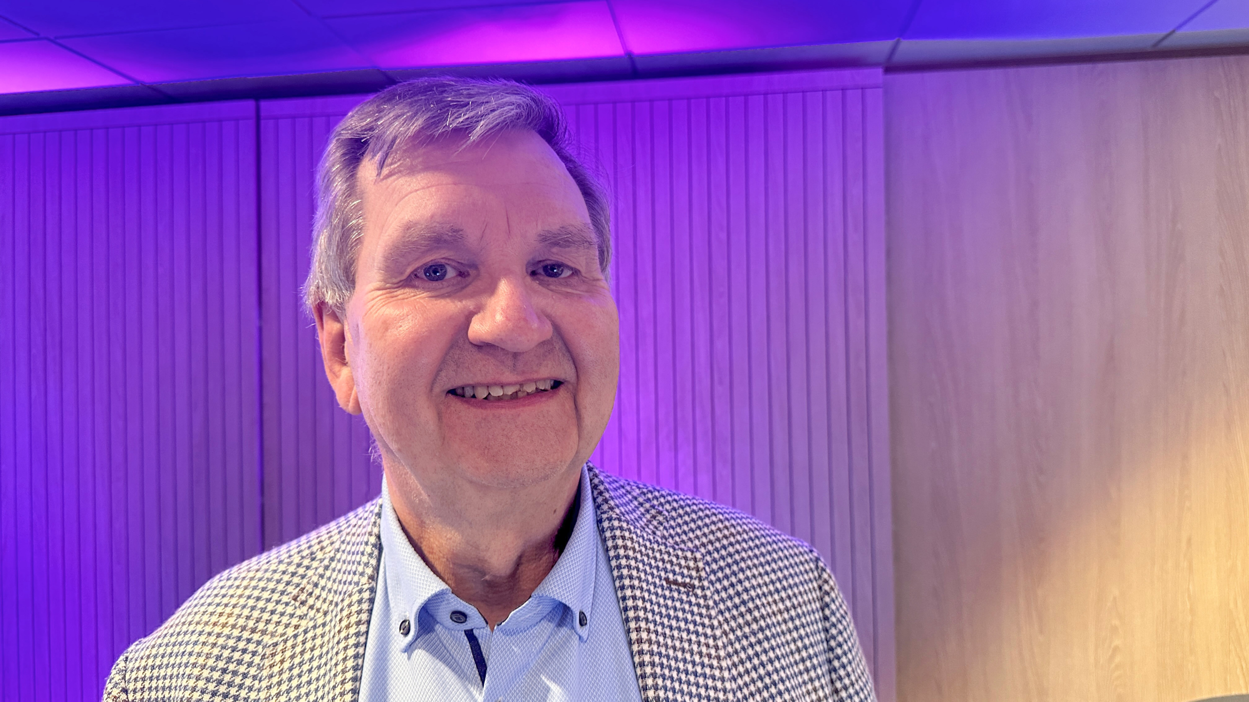 Man in een geruit jasje glimlacht voor een houten wand, met paars verlichte plafondpanelen op de achtergrond.