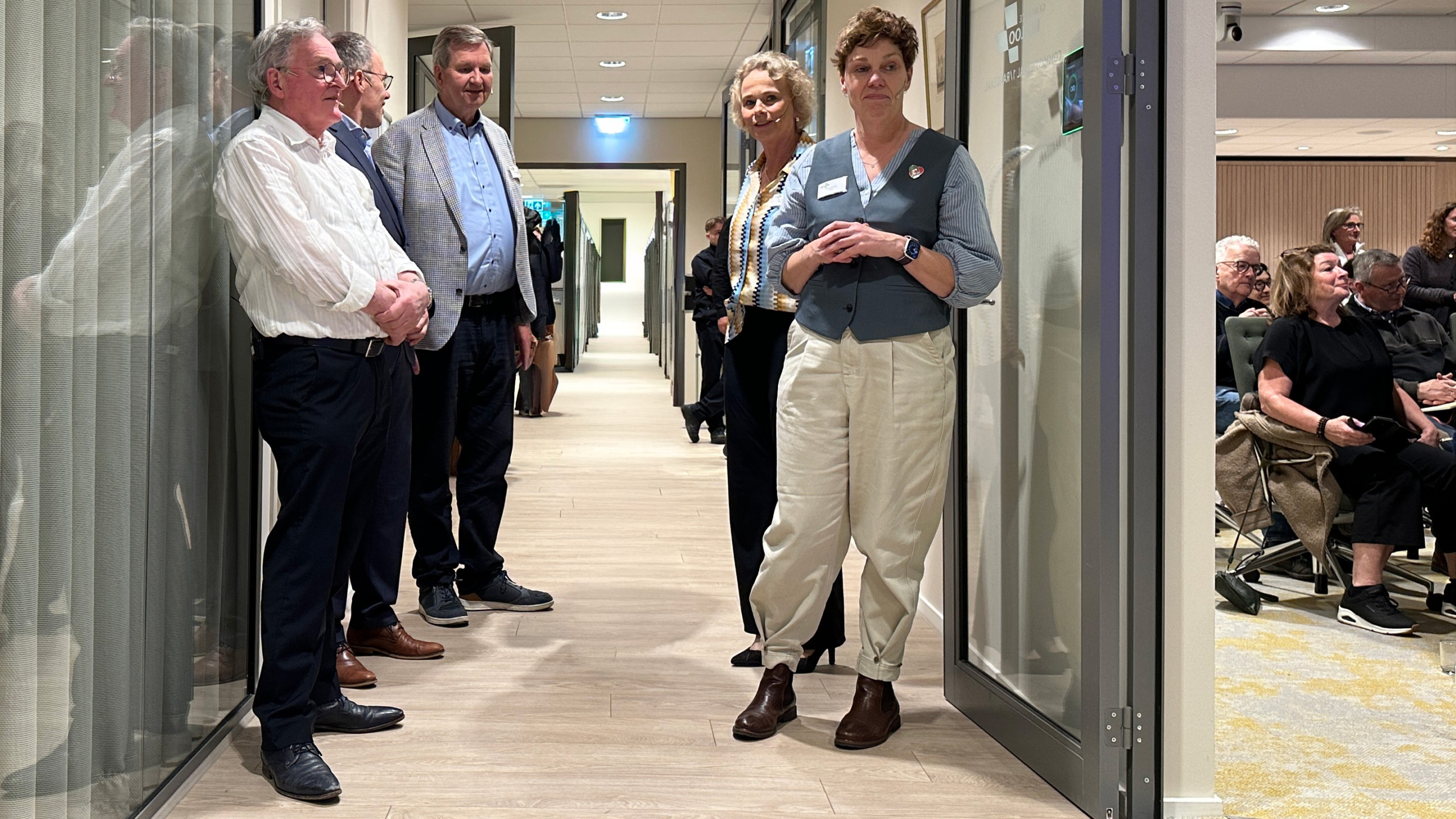 Groep mensen staat in een gang van een gebouw; enkele zitten in een aangrenzende ruimte met stoelen.