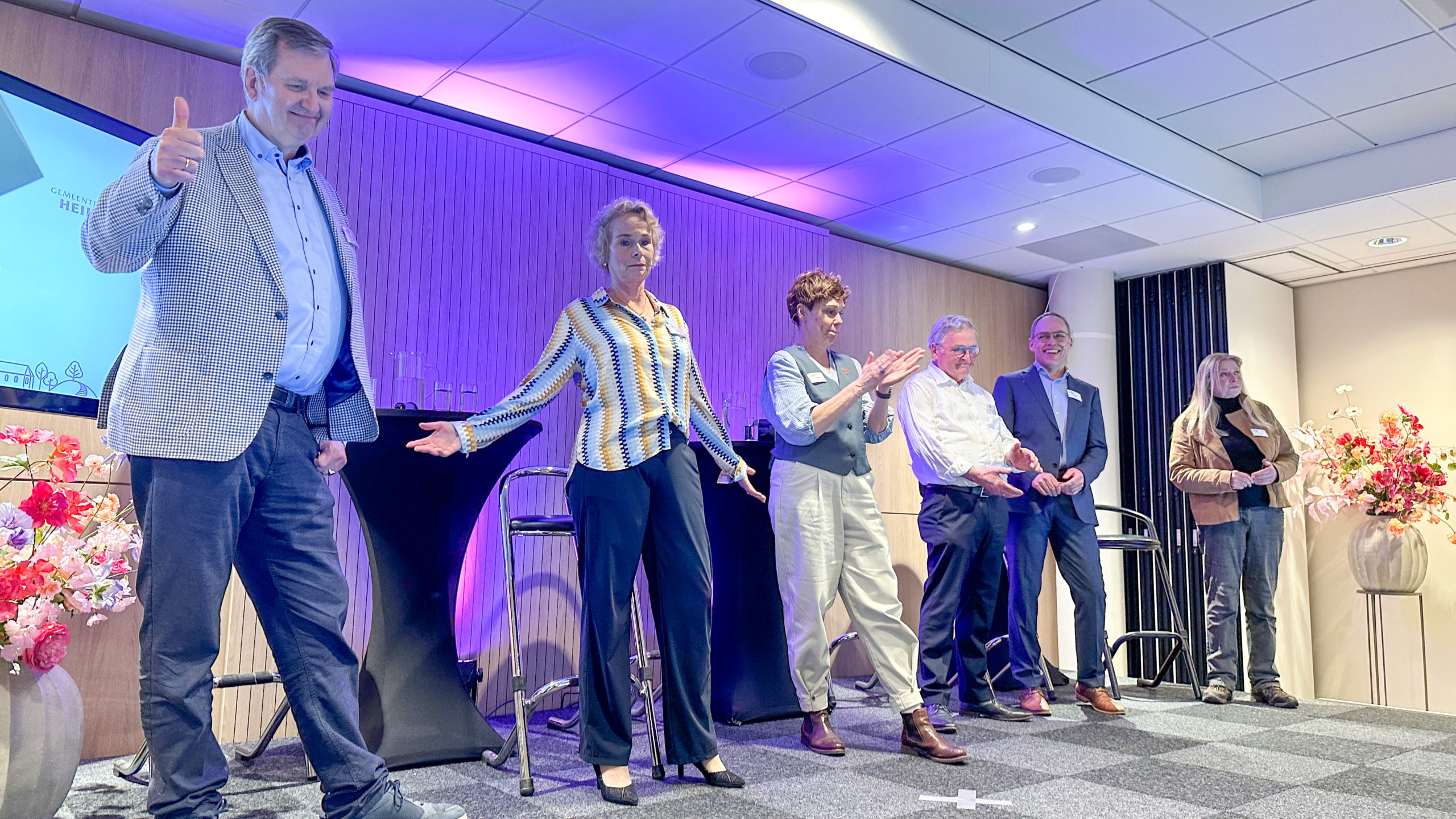Zes mensen staan naast elkaar op een podium met bloemen en een scherm op de achtergrond. Eén persoon steekt een duim omhoog.